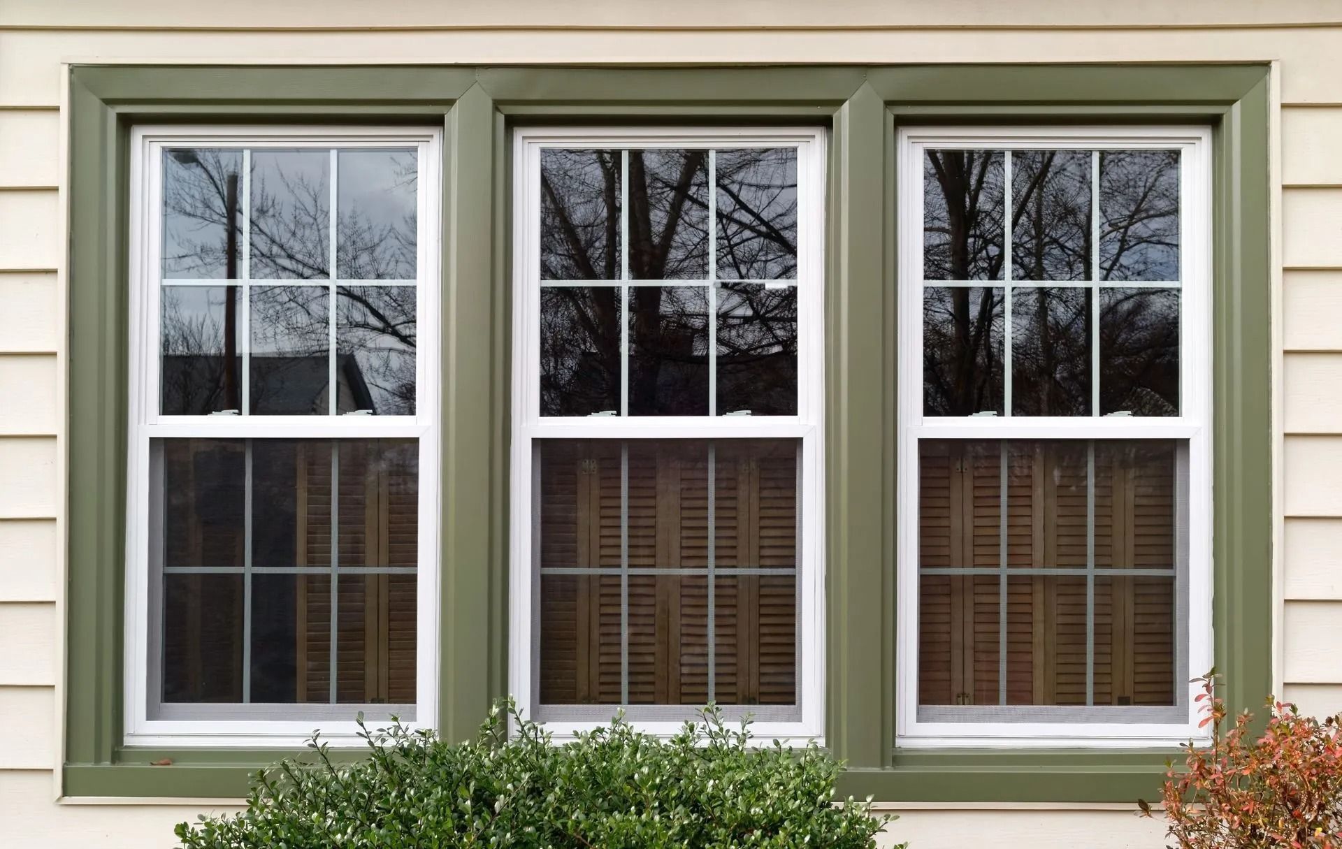 Three white-framed windows with green trim on a cream-colored house. Reflective glass shows trees and shutters.