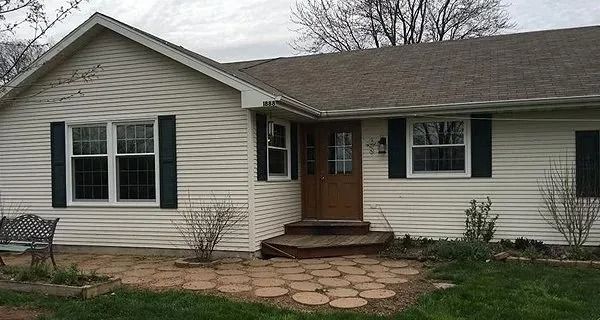 Tan-sided ranch-style house with dark shutters, brown door, and stone pathway. Overcast sky.