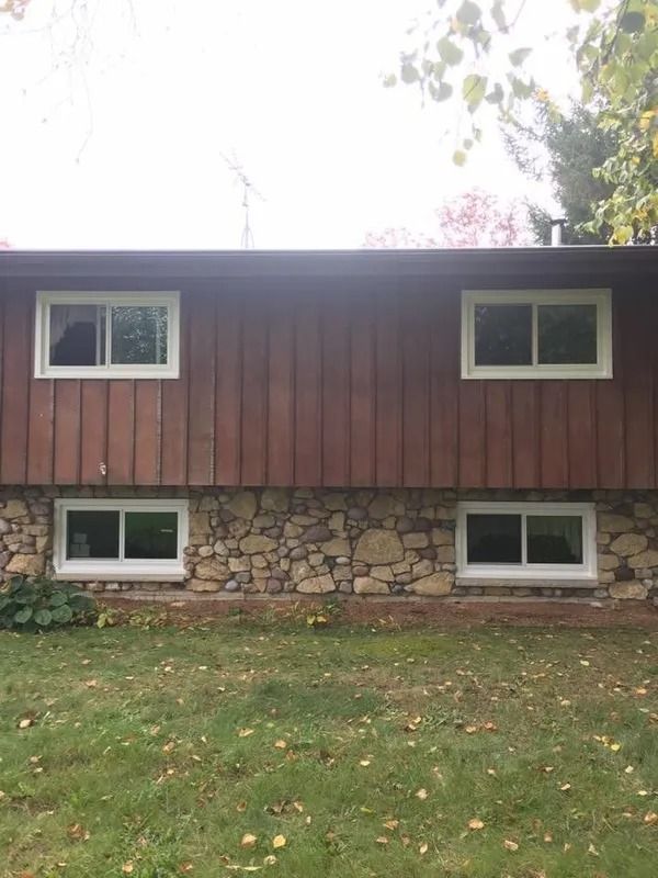 Two-story house exterior. Brown siding above stone foundation with four white-framed windows each level. Green grass.