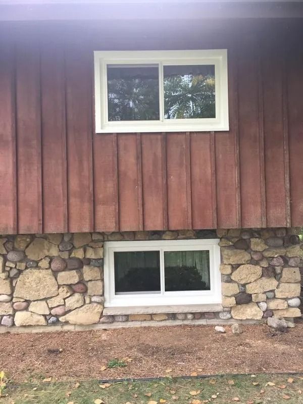 Exterior of a house with two white-framed sliding windows set into brown siding and a stone foundation.
