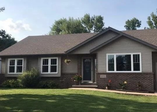Ranch-style house with gray siding, brick accents, and white-framed windows, against a blue sky.