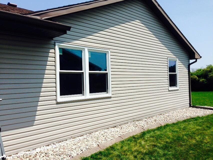 Tan vinyl siding on a house with white-framed windows, a dark roof, and a gravel border.