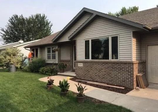 House with tan siding, brown brick, and dark gray trim, surrounded by a green lawn and plants.