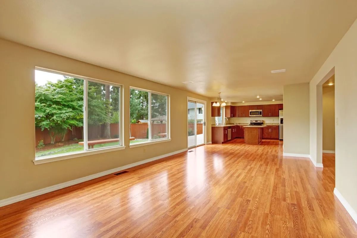 Empty room with wood floors and windows overlooking a backyard, leading to a kitchen.
