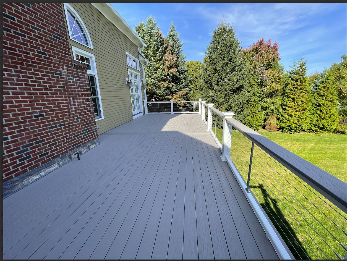 Gray deck with white railing next to a brick building and green lawn with trees.