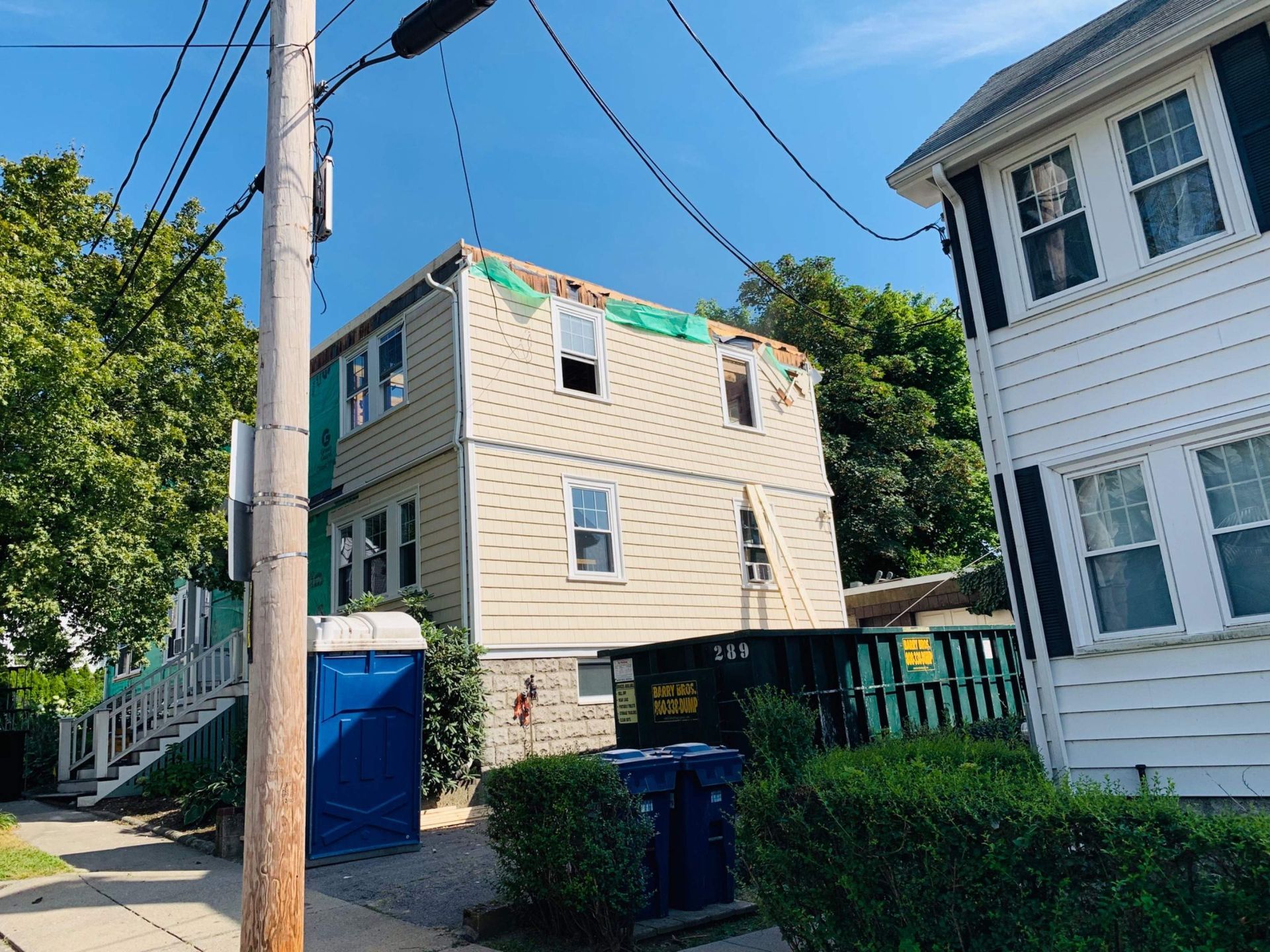 A two-story building with a damaged roof, a blue portable toilet, and green bushes.