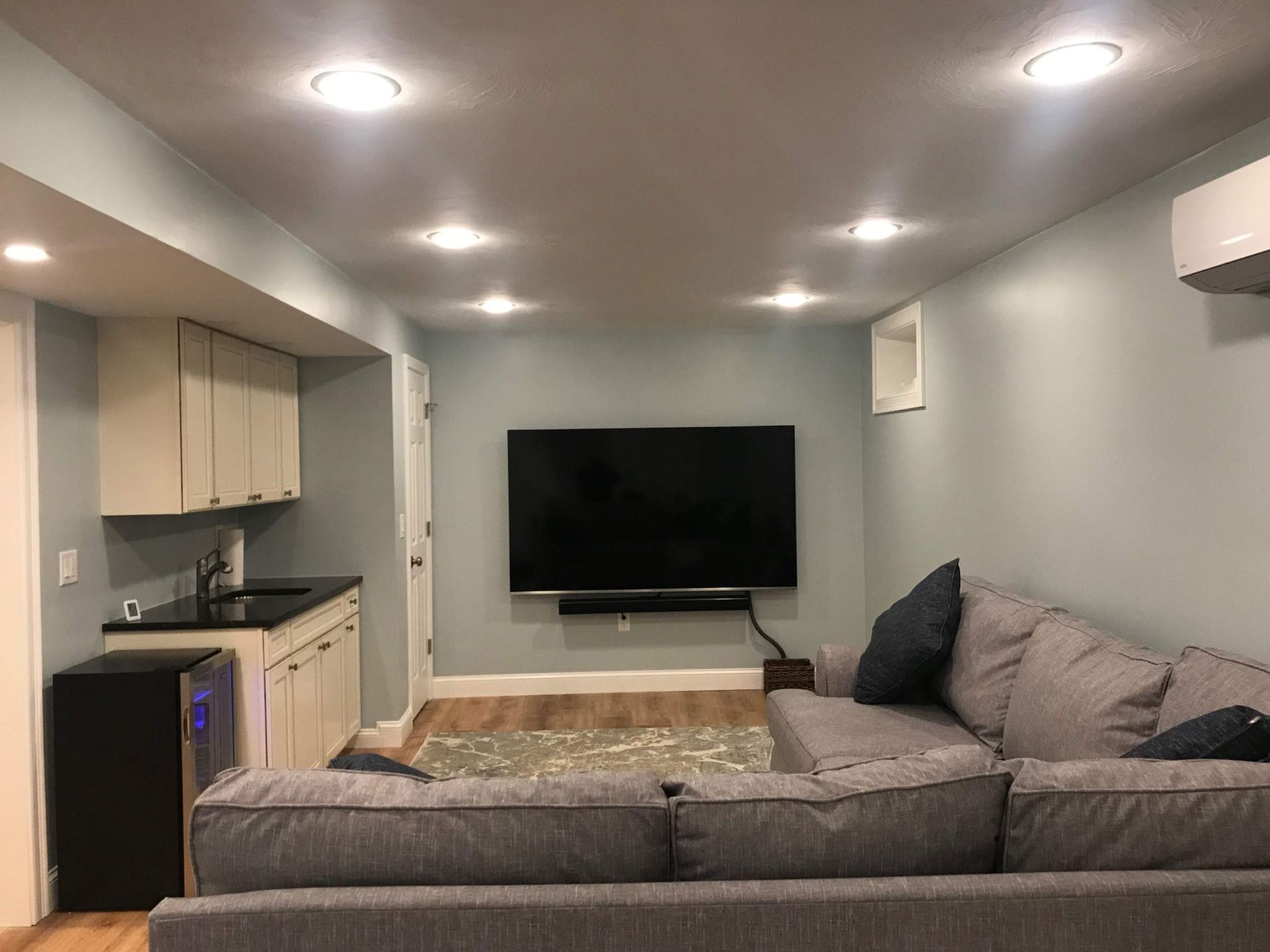 A finished basement with light blue walls, a gray sectional sofa, a TV, and a kitchenette.