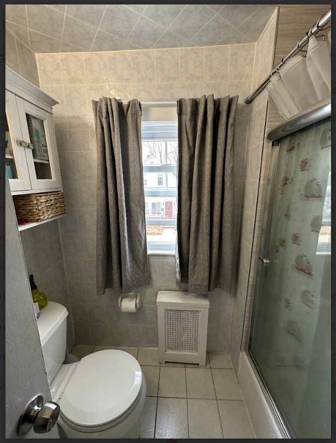 Small bathroom with toilet, shower, and window with curtains. Gray tile walls and a cabinet.