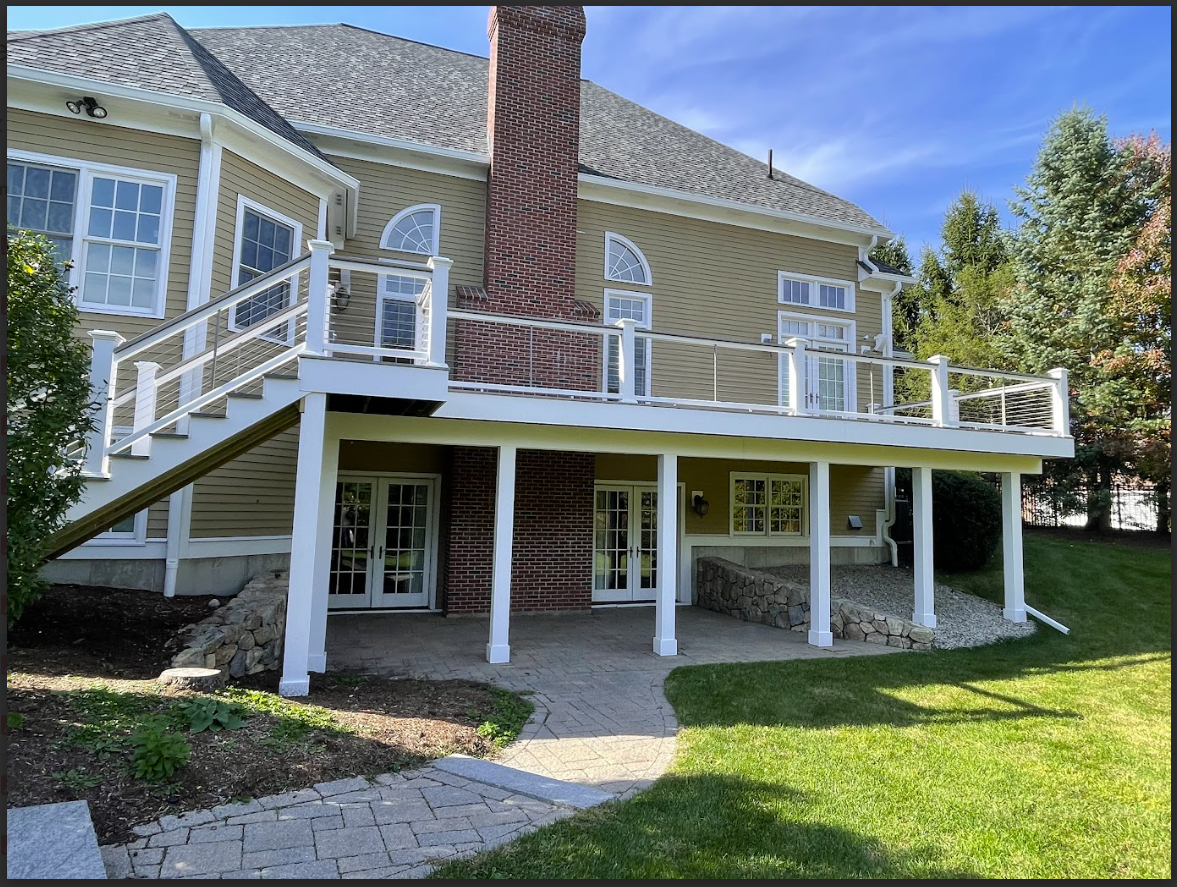 Back view of a light beige house with white deck, brick chimney, and stone patio.
