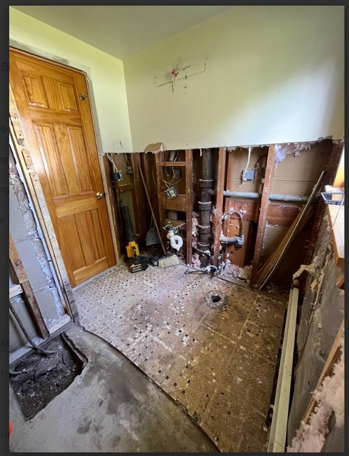 Bathroom renovation in progress. Walls torn open, exposing plumbing. Door on the left, debris on the floor.