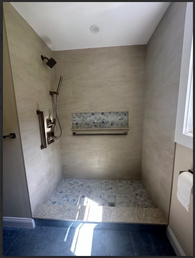 Accessible shower with pebble floor, beige tile walls, bronze fixtures, and built-in shelf.