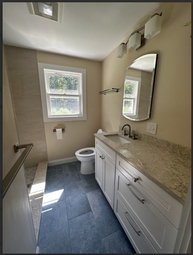 Bathroom with blue tile floor, white vanity, toilet, window, mirror, and light fixture.