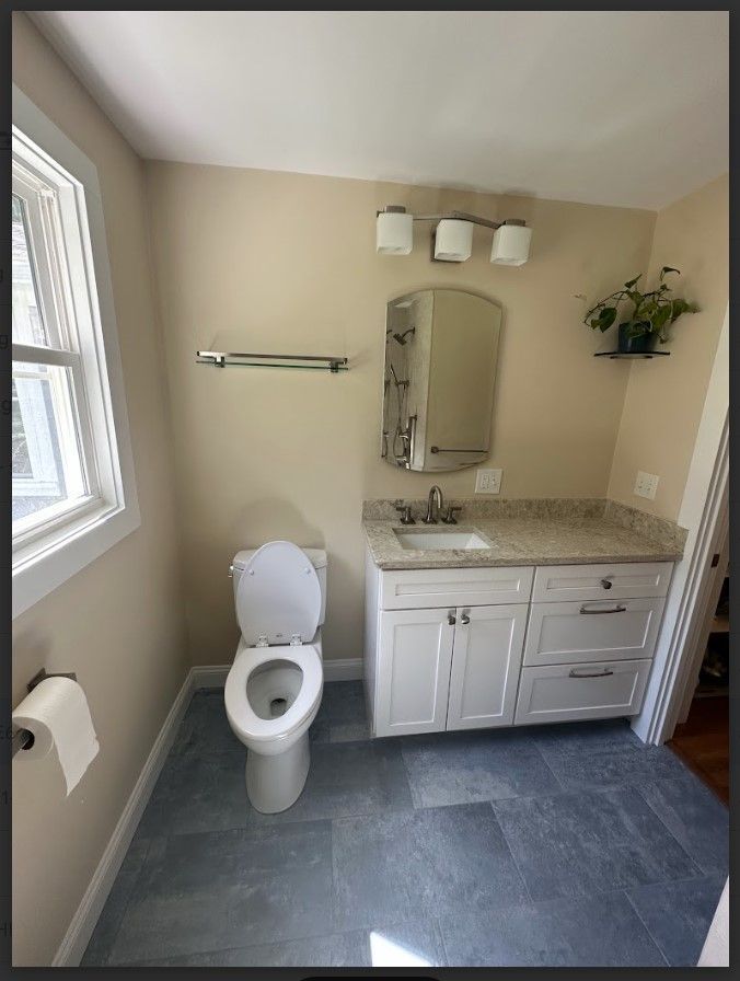 Bathroom with a toilet, vanity, and window. Blue tile floor, light beige walls, and white cabinets.