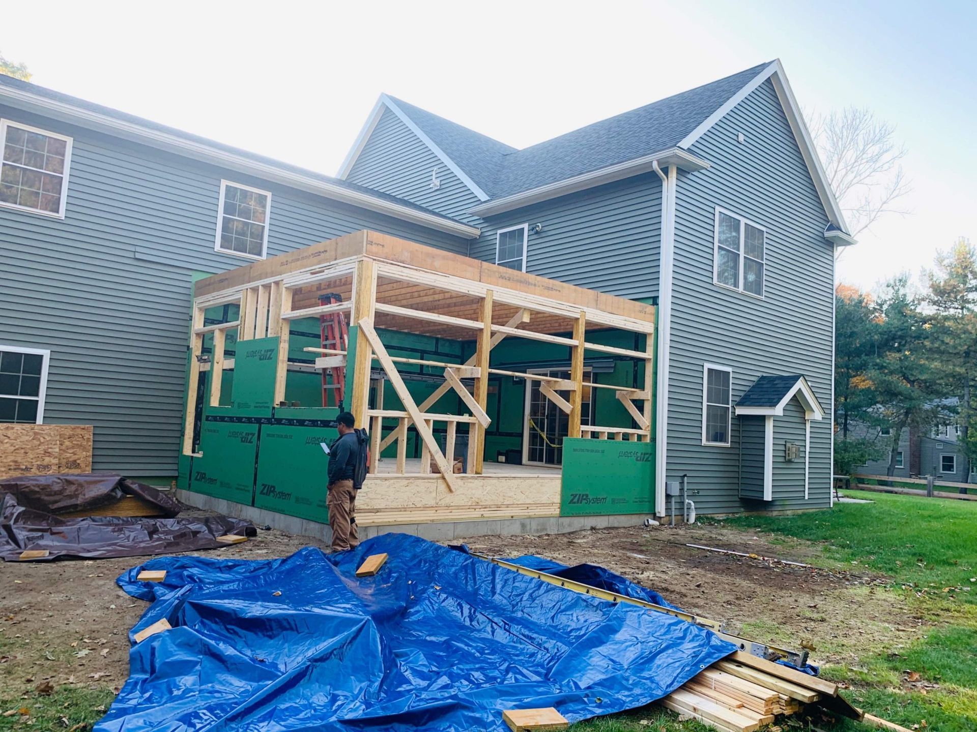 Construction of a room addition on a gray house. A person stands near framing and green sheathing.
