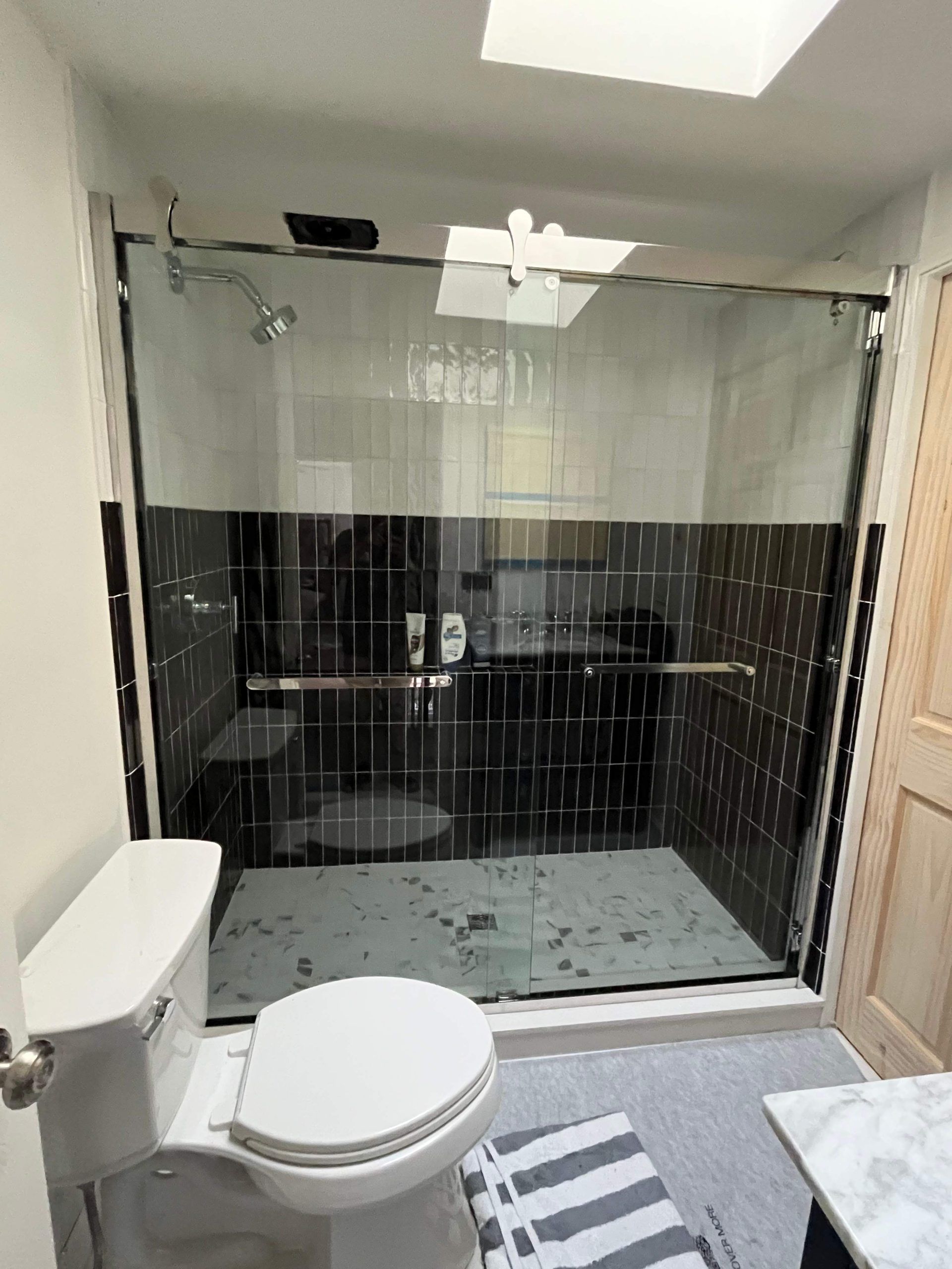Bathroom with black and white tiled shower, glass door, toilet, and gray rug.