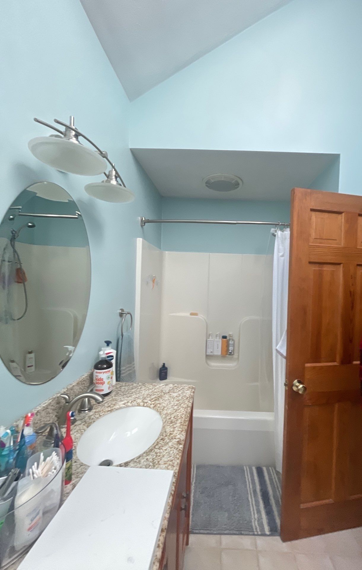 Bathroom with light blue walls, vanity, oval mirror, shower, and open wooden door.