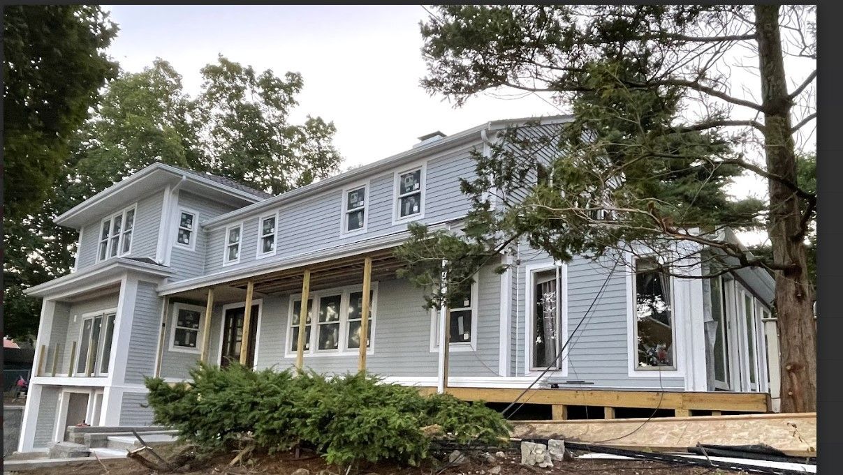 Two-story light blue house under construction with a porch and surrounding trees.
