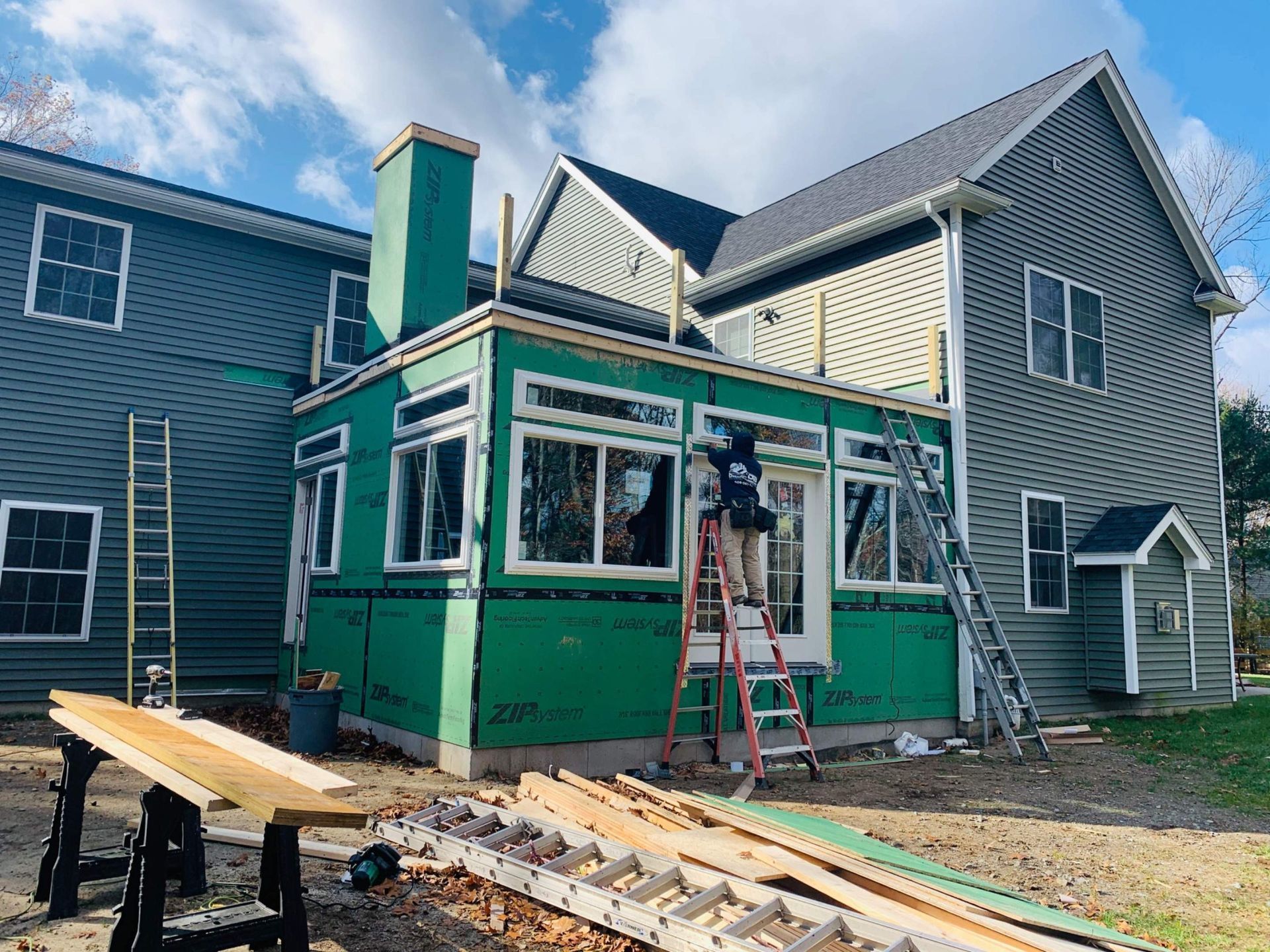 Construction of a sunroom addition with windows and green siding attached to a gray house. 