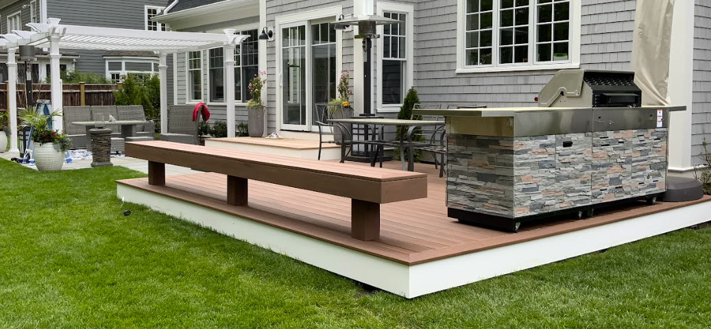 A backyard deck with a built-in grill, bench seating, and a pergola. Green grass surrounds the deck.