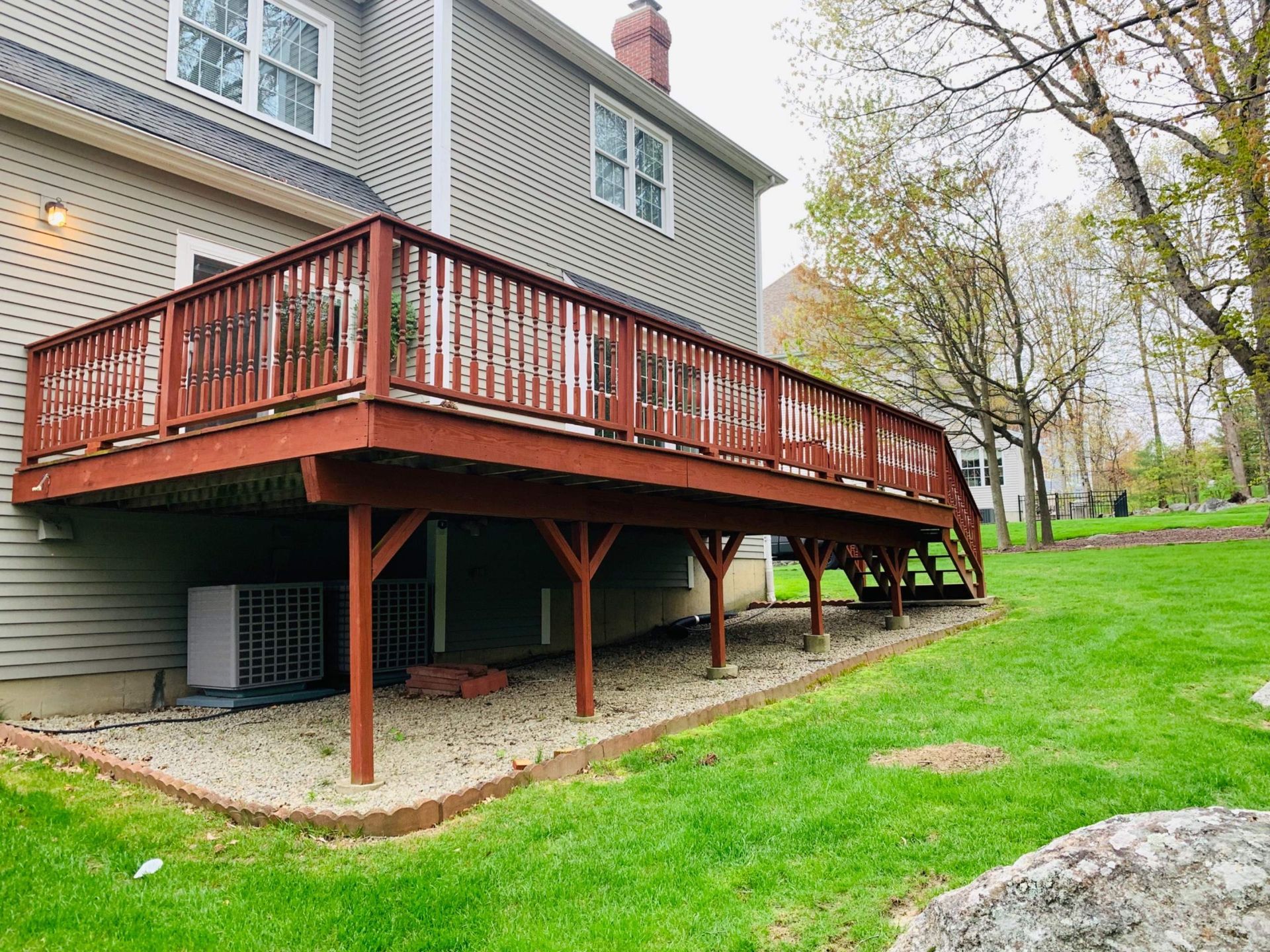 Red wooden deck attached to a gray house, supported by posts. Gravel ground and green lawn surround it.