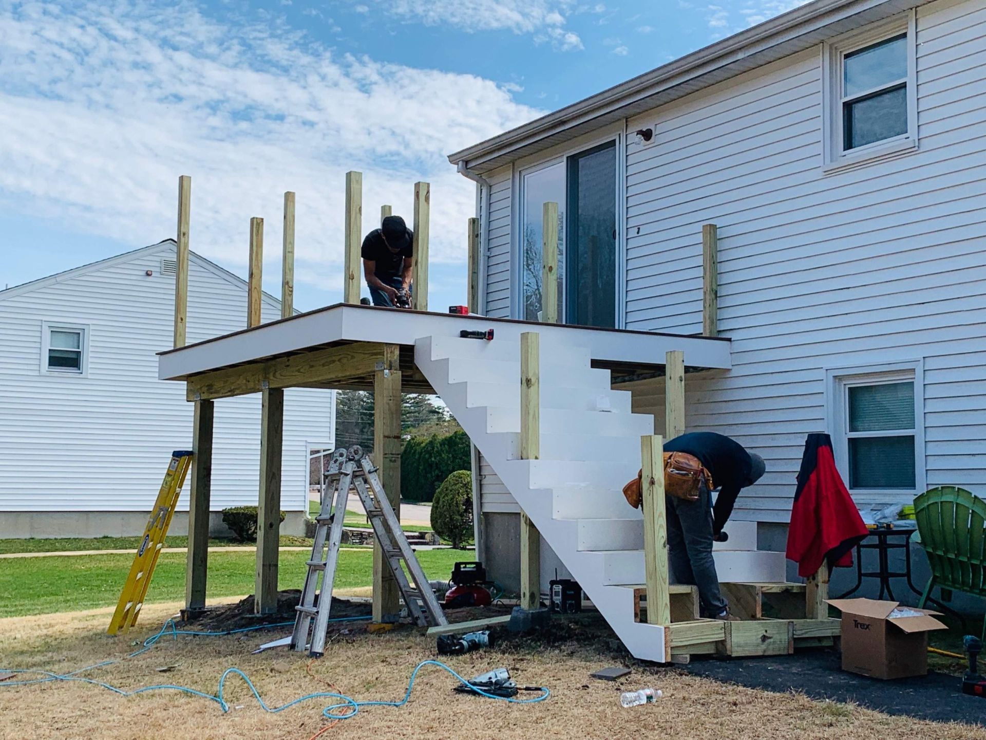 Two people building a deck attached to a white house. Wooden posts and stairs are visible.