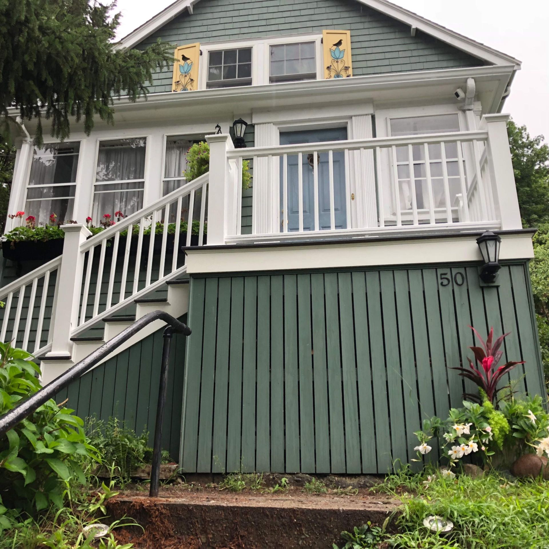 Green house with white railing, steps, and door.  Flowers and plants in front. Number 50 on the side.