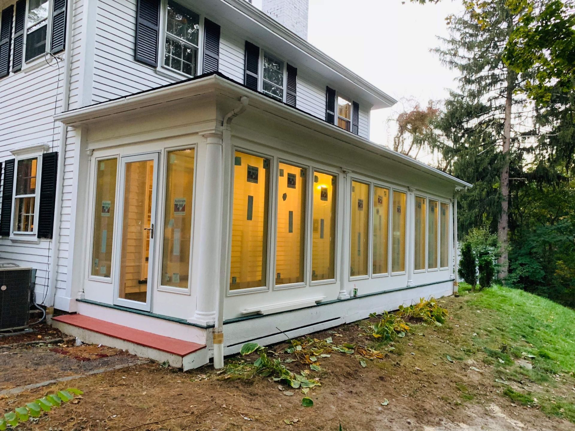 White house with a sunroom addition, featuring many windows and a red-painted platform. Green grass and trees surround it.