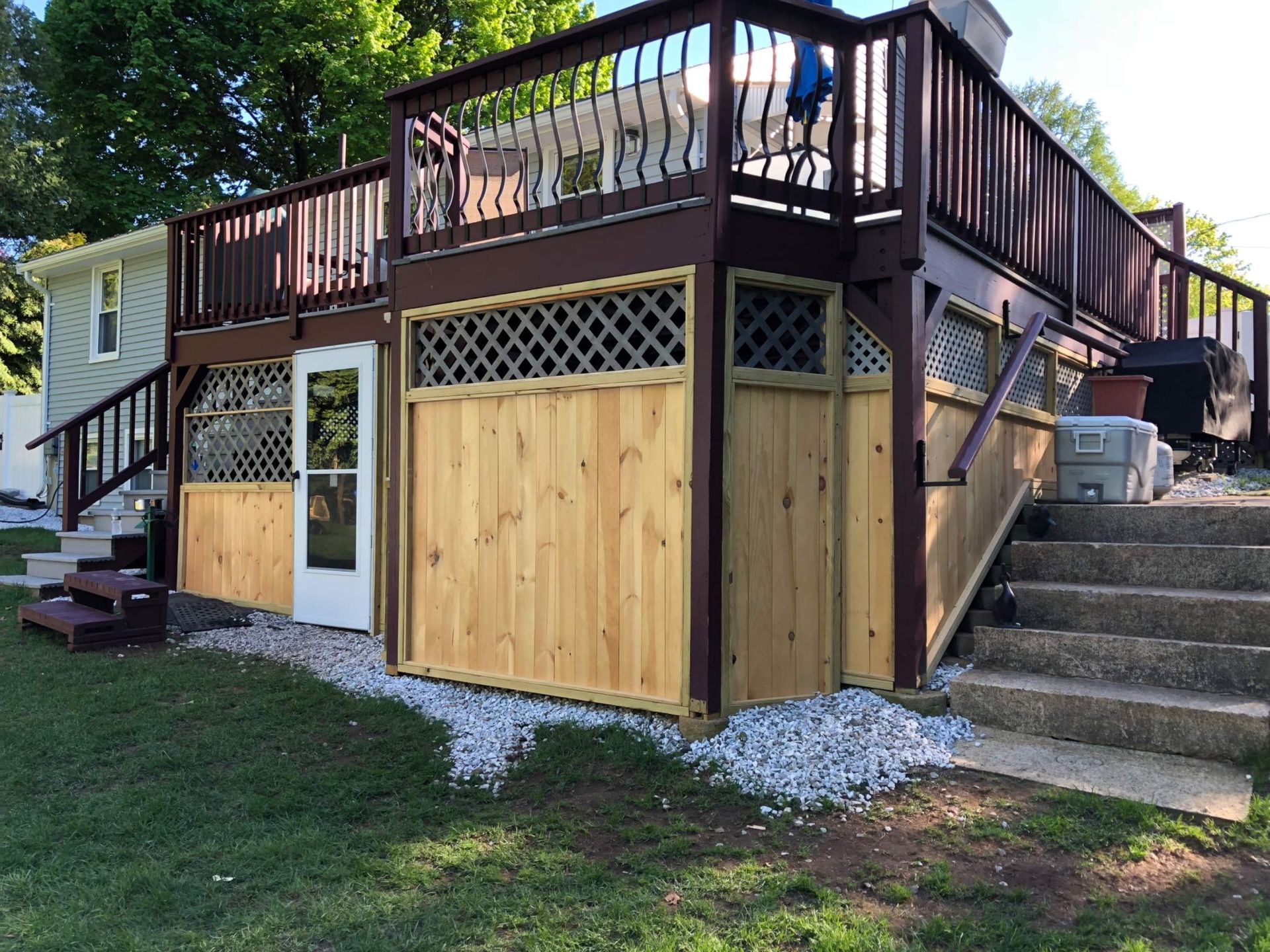Two-level wooden deck with lattice and natural wood panels, steps down to a concrete path and a grassy lawn.