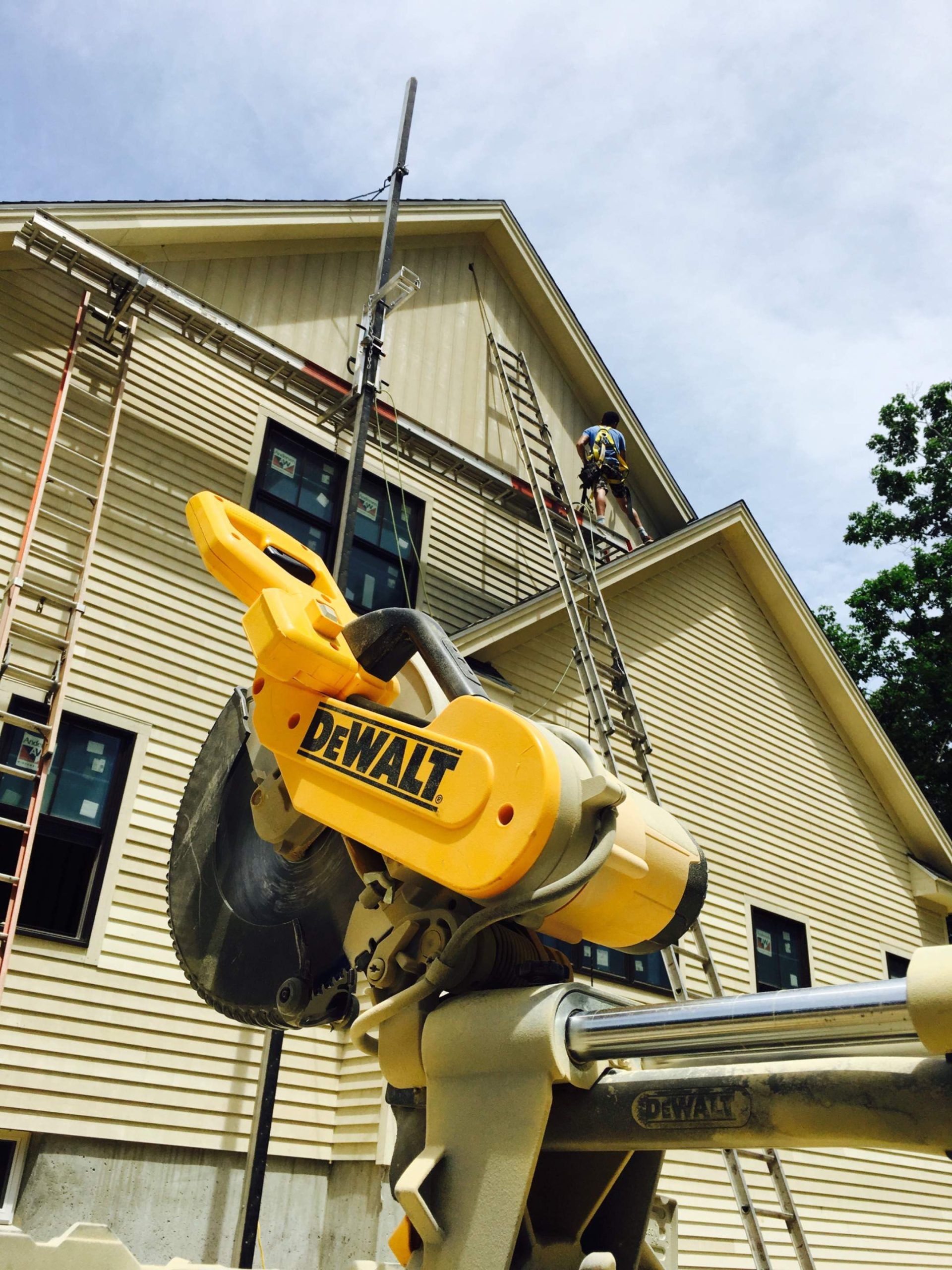 Yellow DeWalt miter saw in front of a house under construction; a worker on a ladder.
