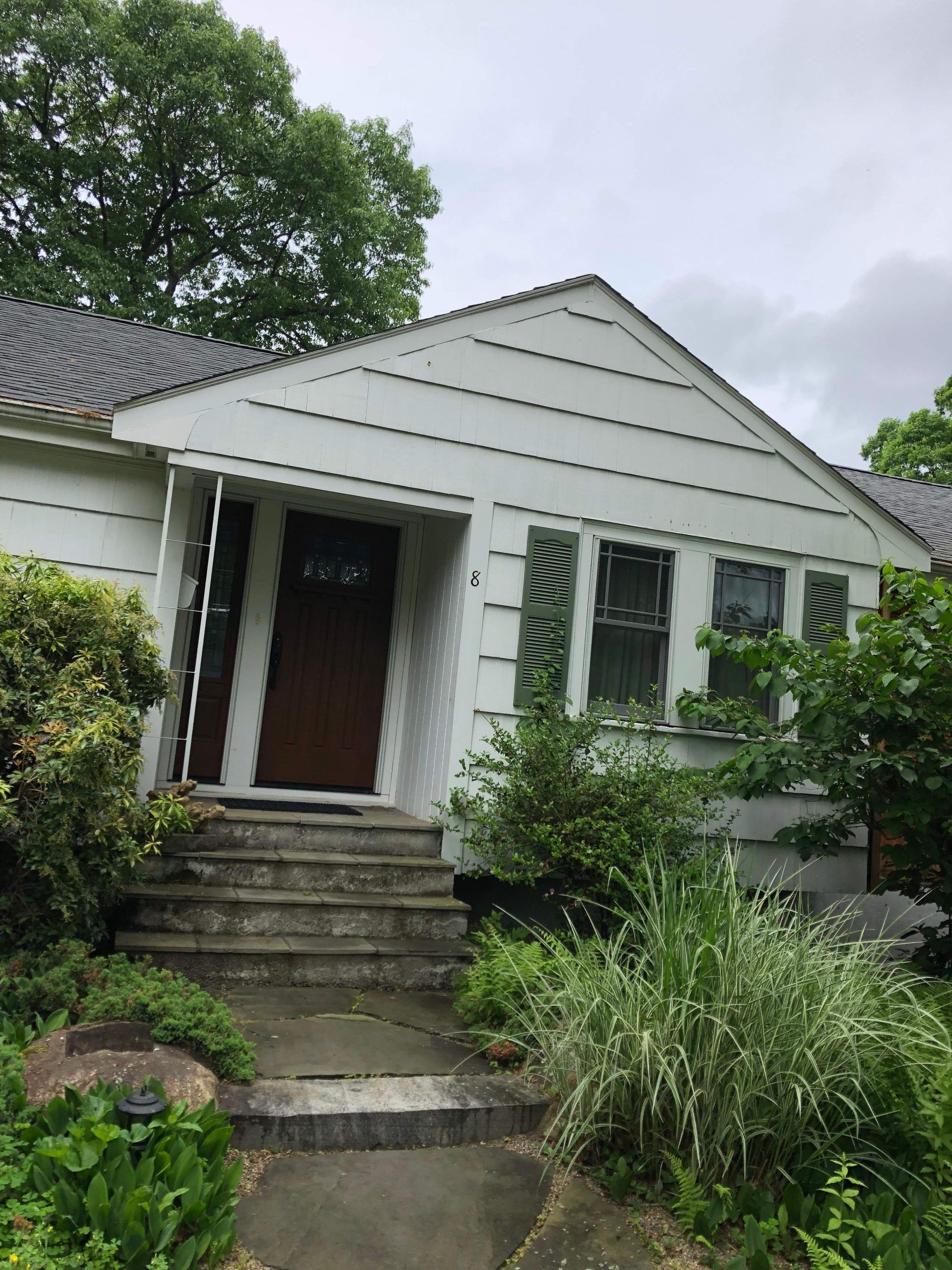 White house with dark door, grey steps, and green shutters. Landscaping includes bushes and tall grass.