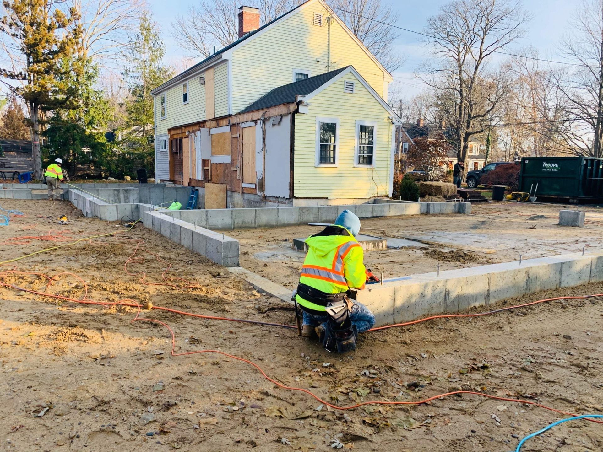 Construction worker kneeling, working on a new foundation. Existing house is in background, yellow with exposed studs.