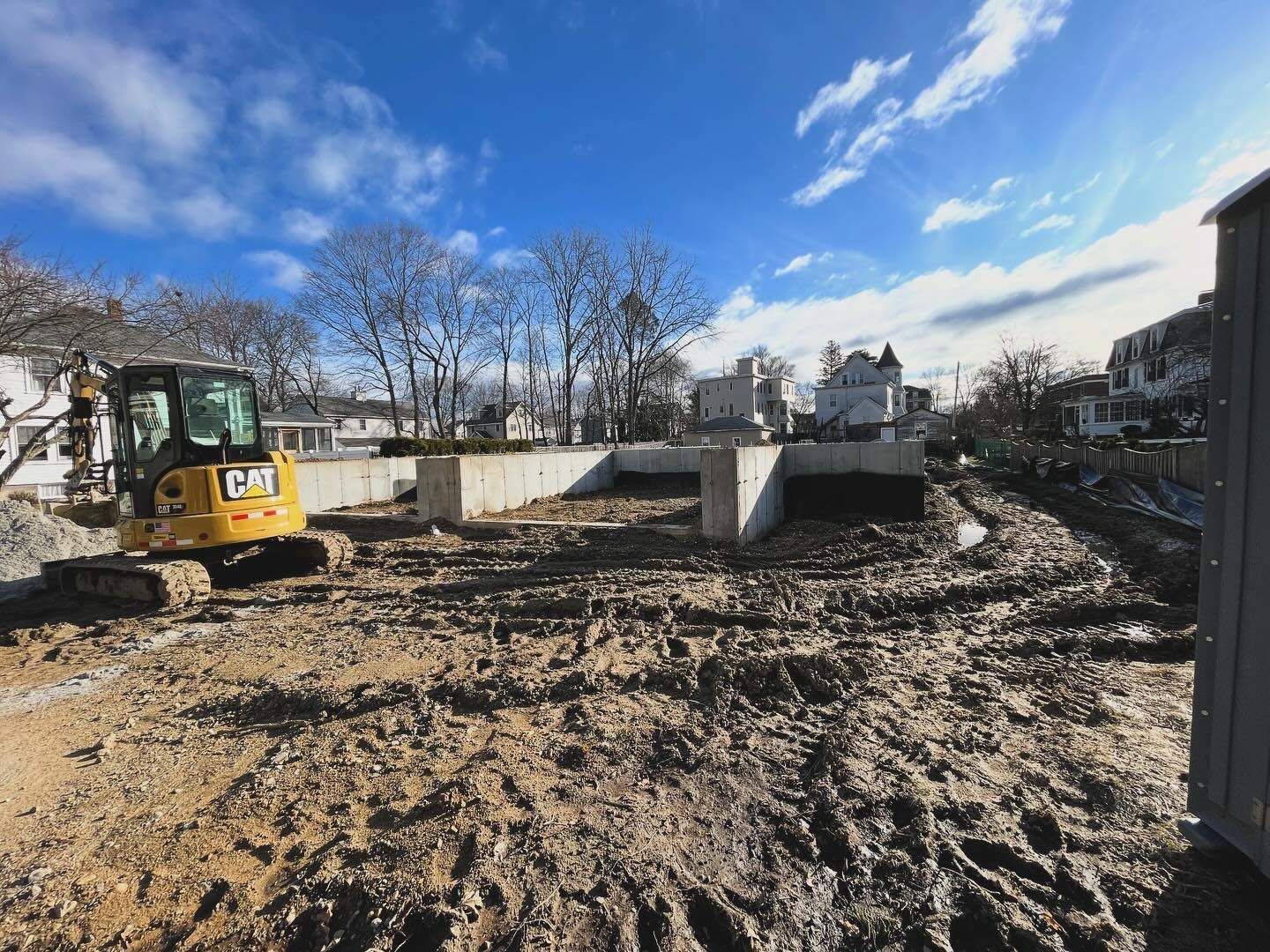 Construction site with a small excavator next to a concrete foundation under a blue sky. Muddy ground.