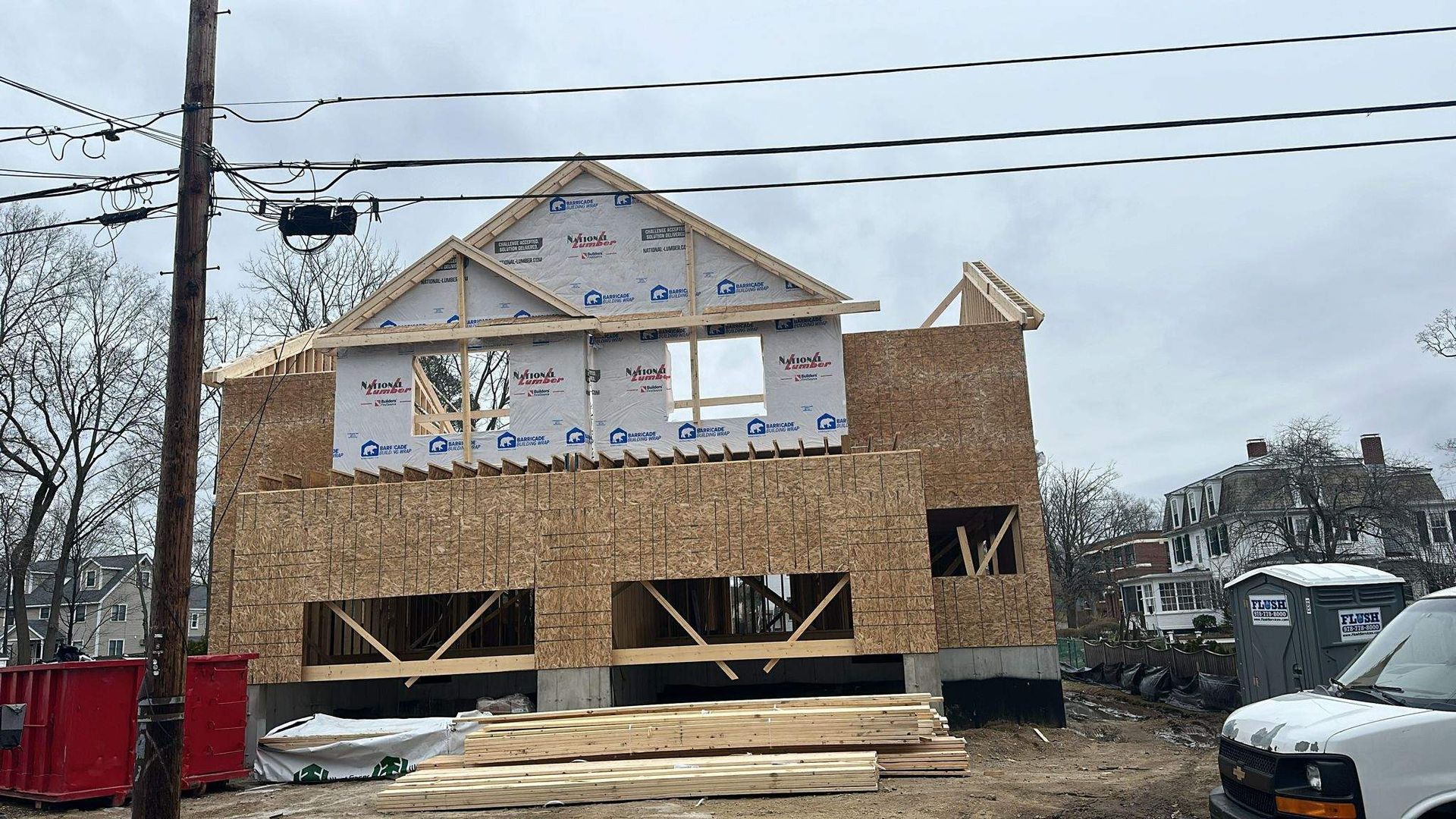 Construction site of a two-story house, wooden frame and plywood siding, under a cloudy sky.