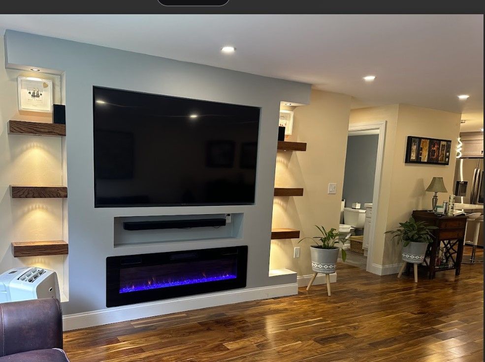 Living room with built-in fireplace and TV, floating shelves with spotlights, light blue accent wall, wooden floors.
