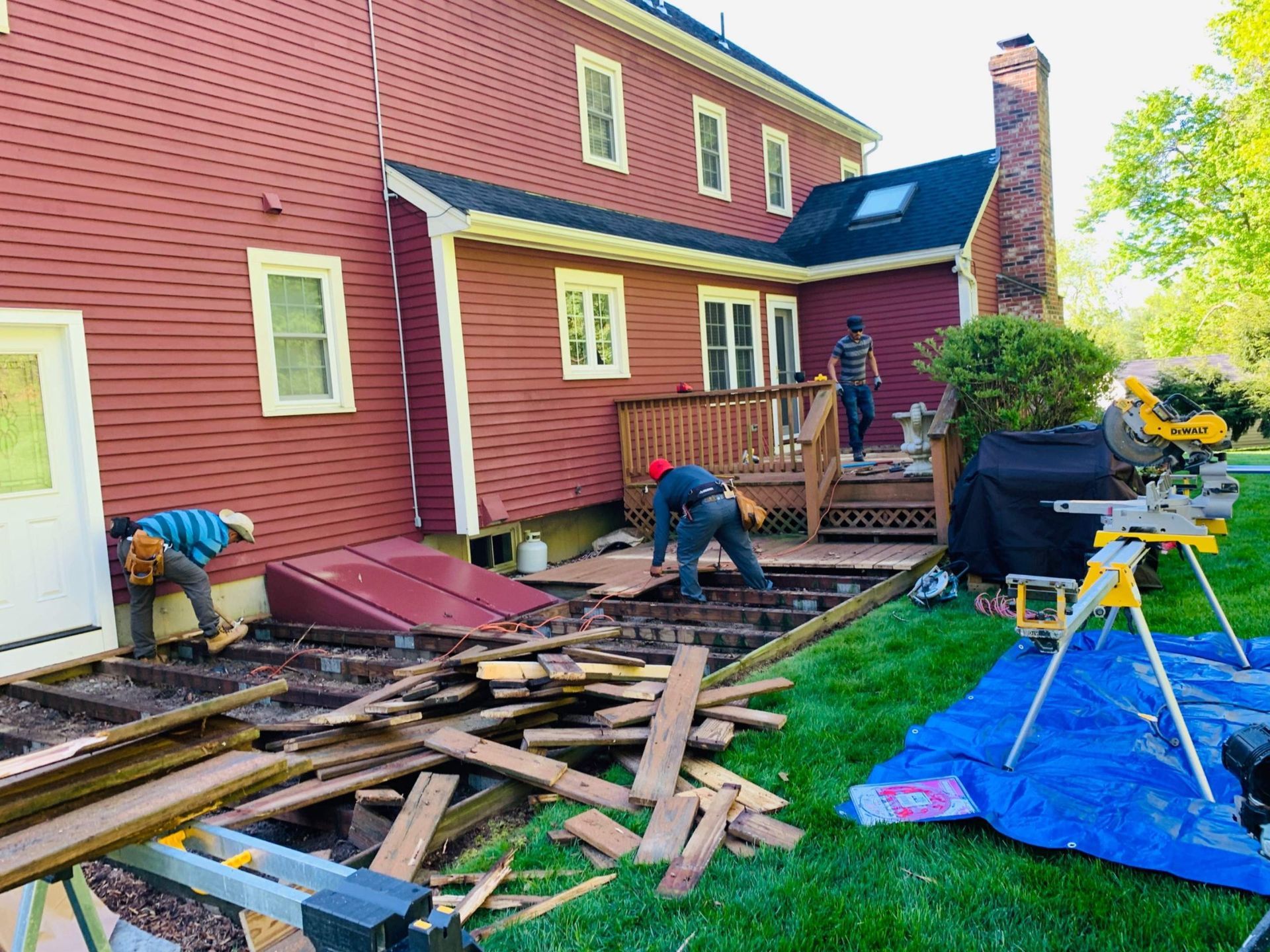 Workers rebuilding a wooden deck next to a red house. Sawing wood and removing old deck boards.