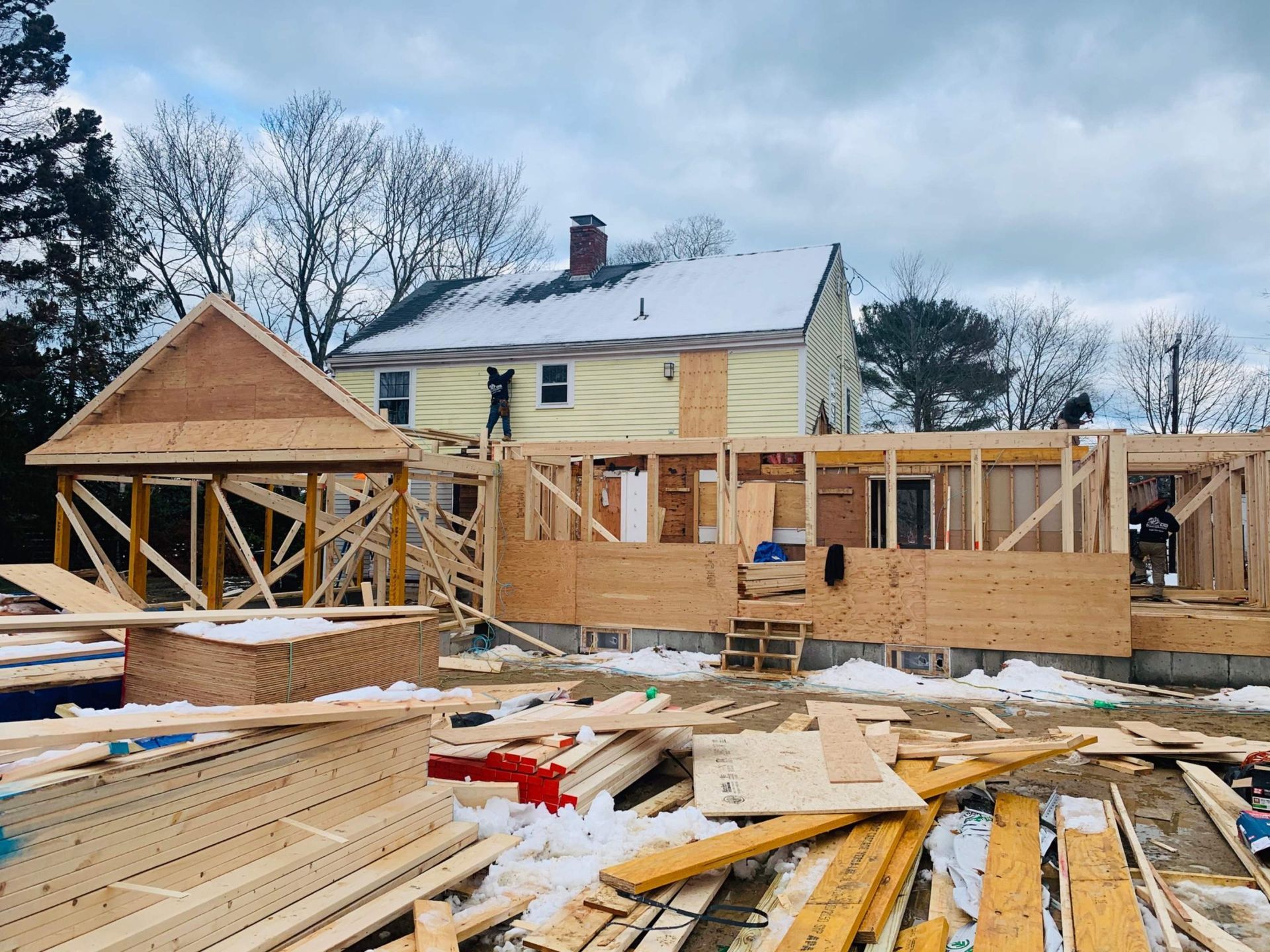 Construction site with wood framing, attached to a yellow house with a snowy roof. Two workers are present.