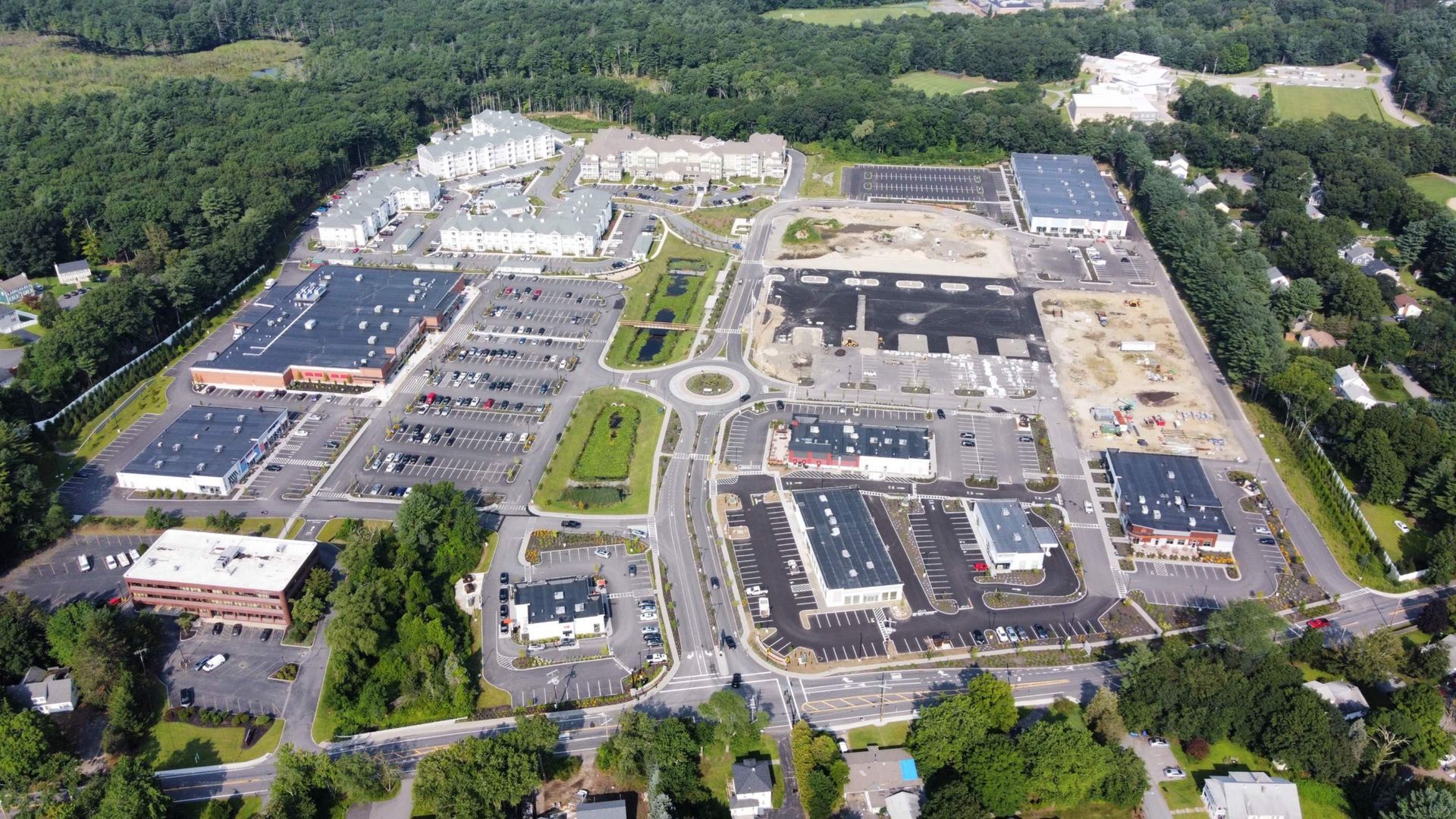 Aerial view of a commercial complex with stores, parking lots, and landscaping, surrounded by trees.