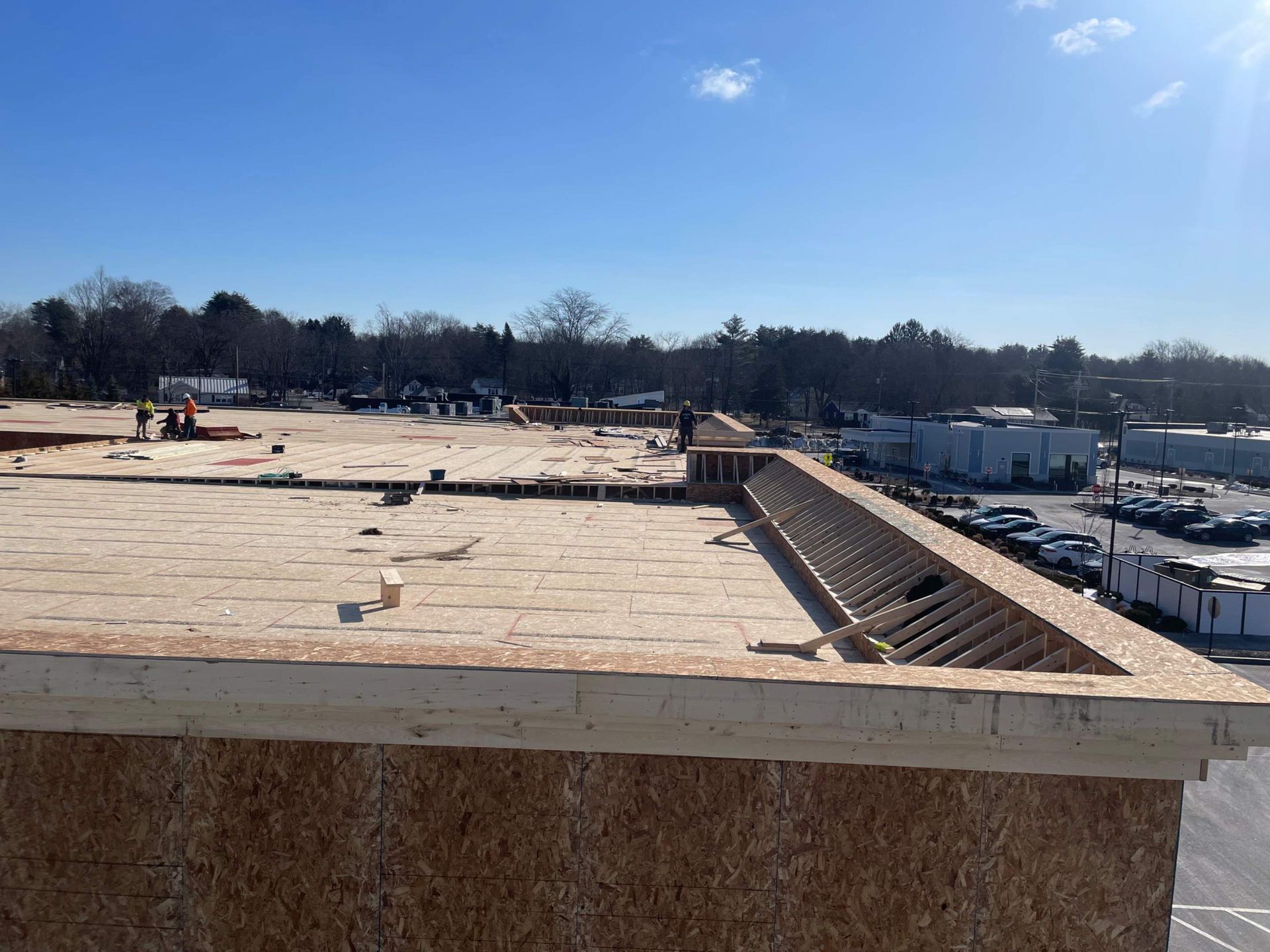 Rooftop construction site under a clear blue sky. Wood framing visible, workers in the distance.