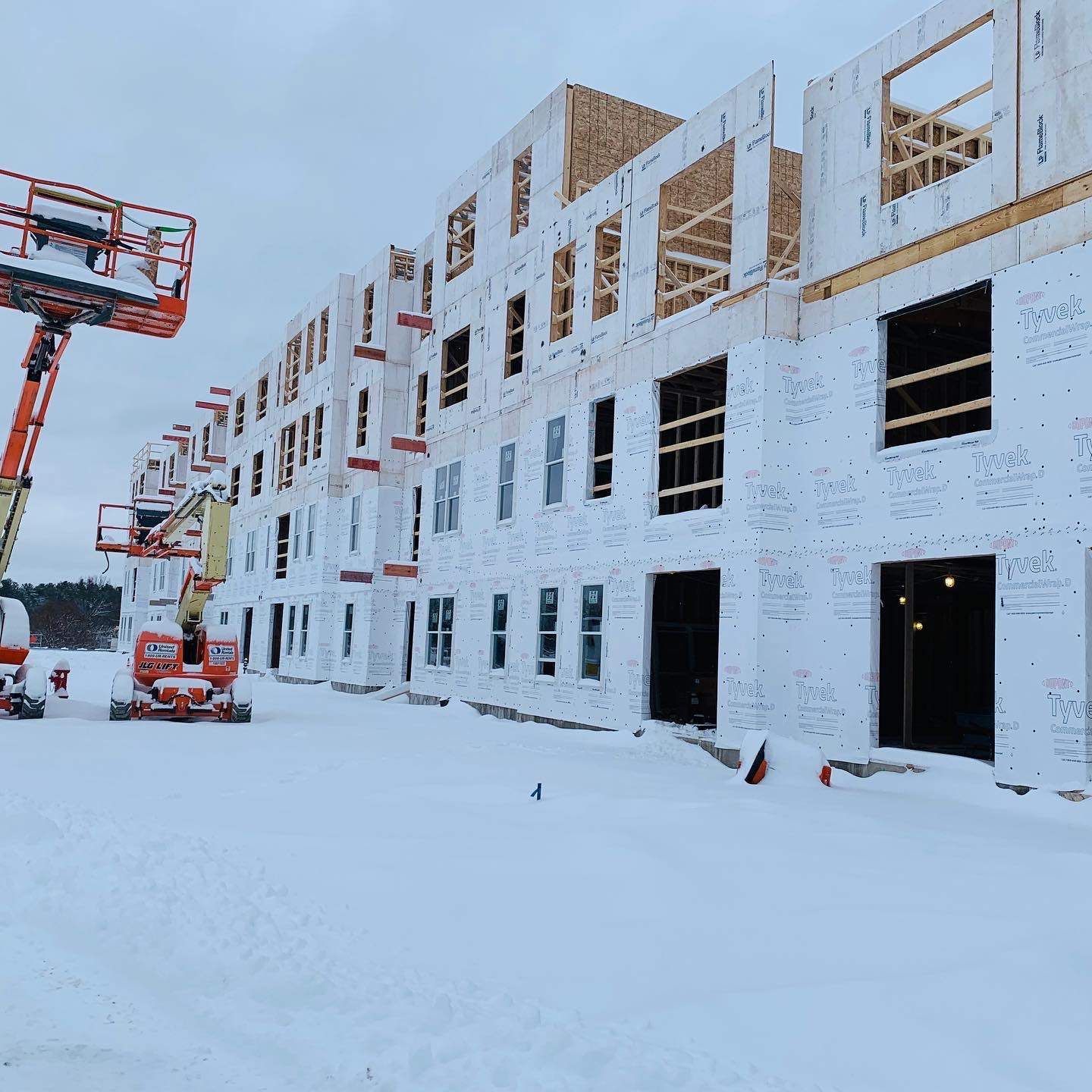 Construction of a multistory building covered in white siding. Snow covers the ground with cherry pickers present.