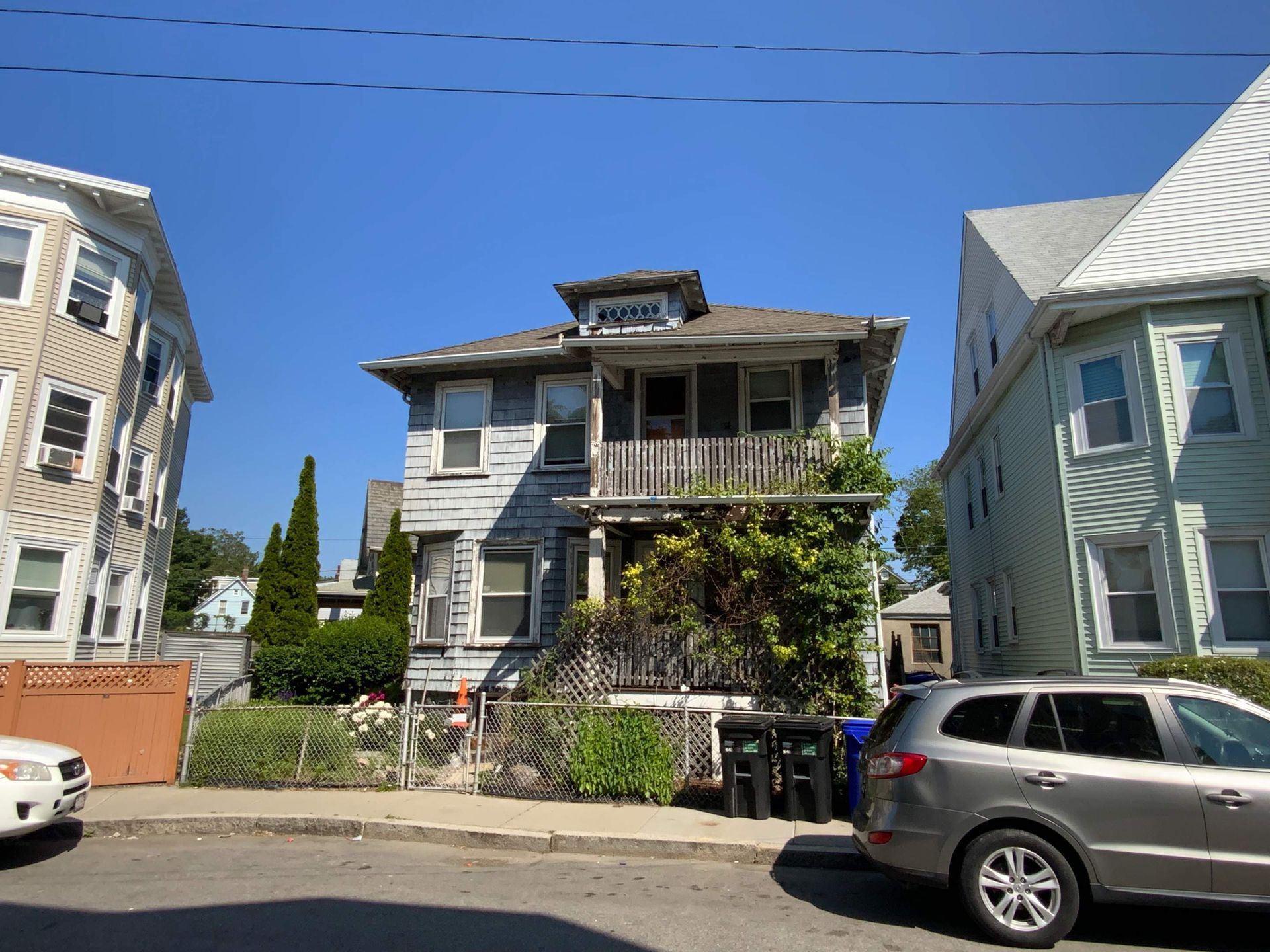 Gray, two-story house with a wooden porch and overgrown bushes. Other houses and a car are also visible.