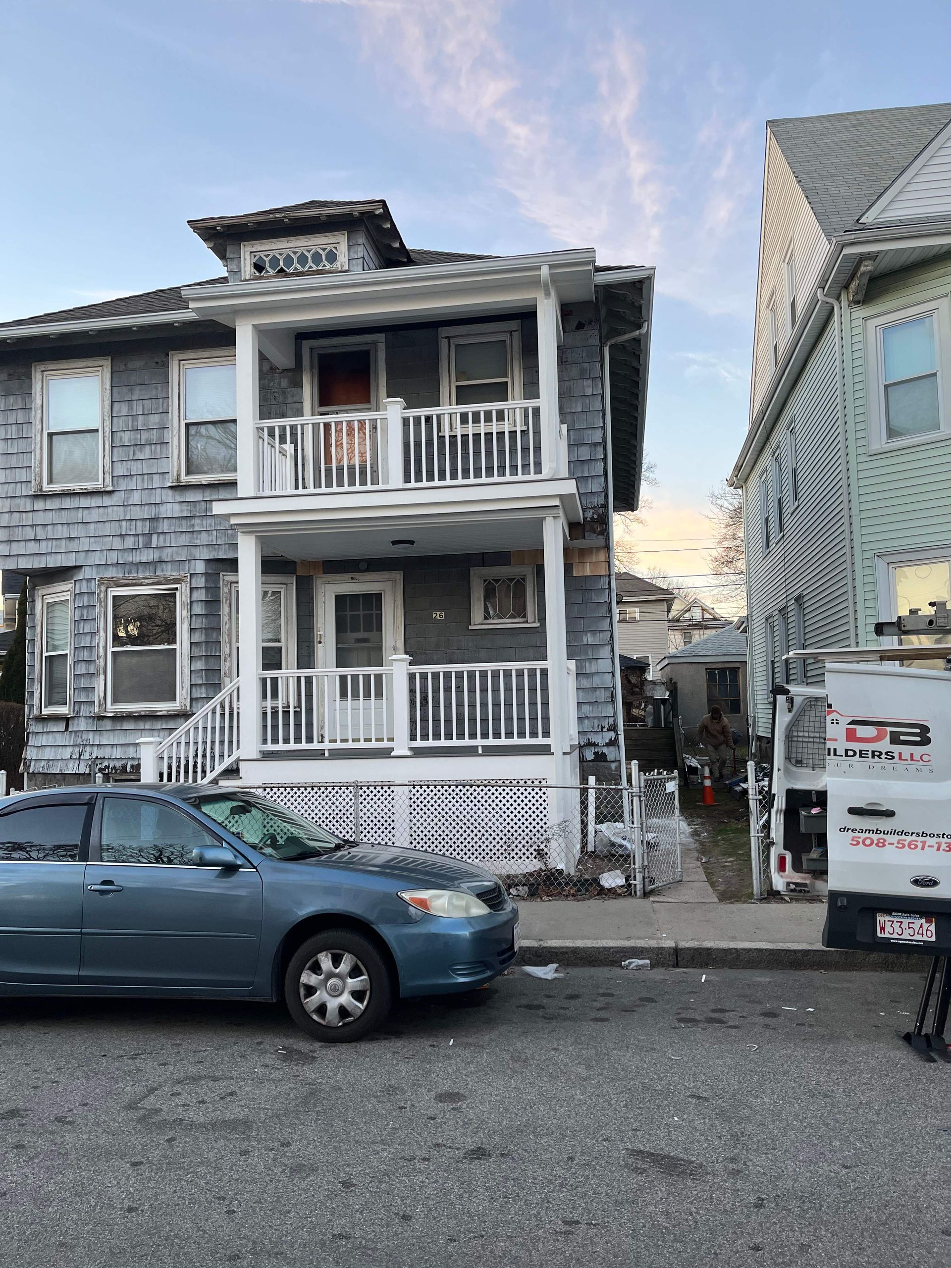 Two-story gray house with a small balcony and a blue car parked on the street.