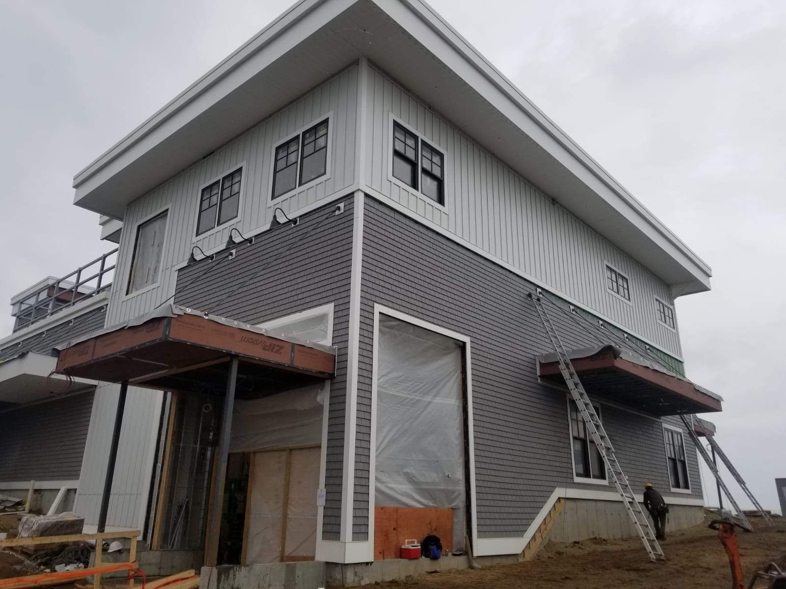 Two-story building under construction with gray shingle and white panel siding. Brown overhangs and black-framed windows.