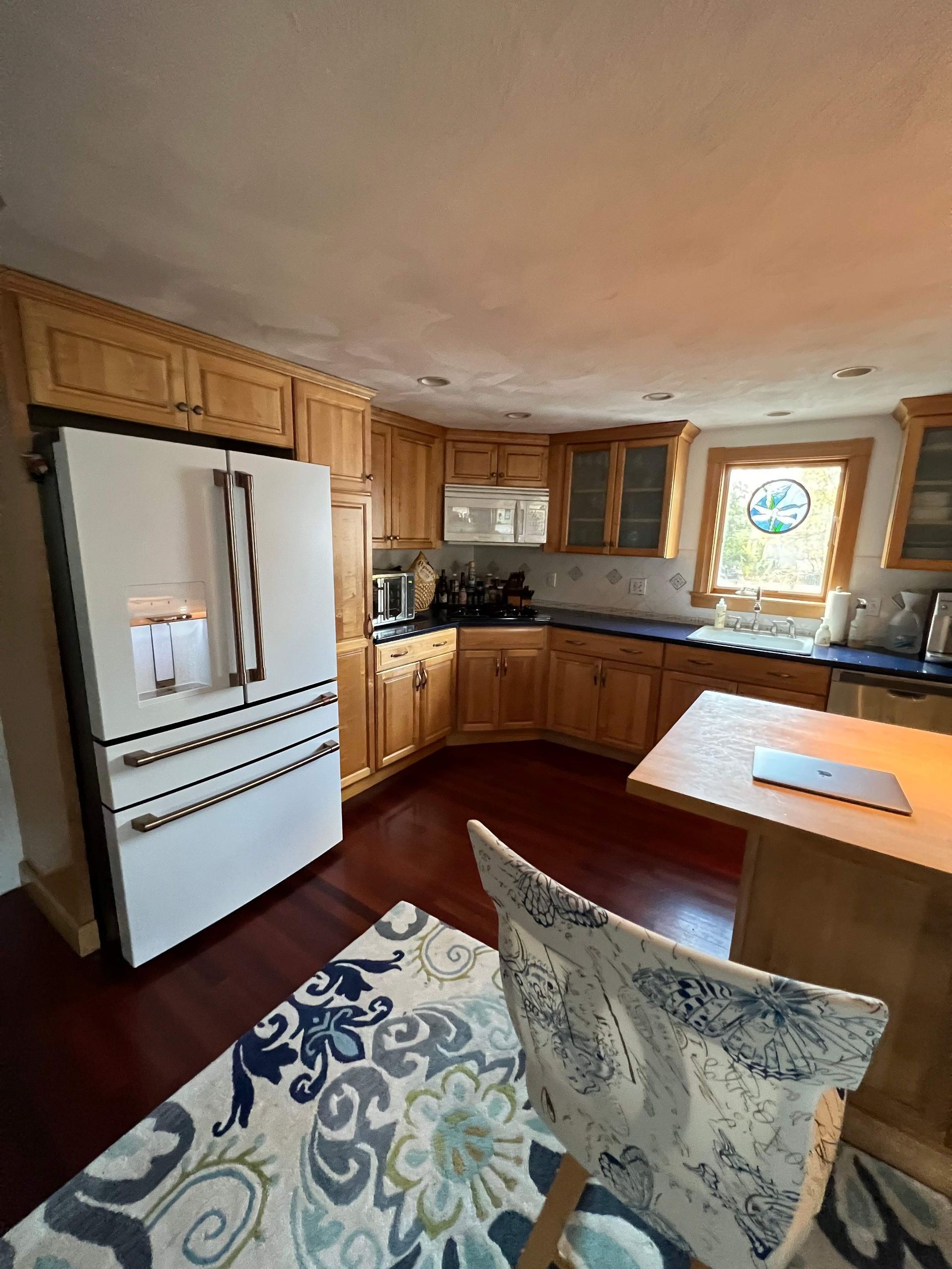 Kitchen with light wood cabinets, white refrigerator, dark red floor, and blue patterned rug.