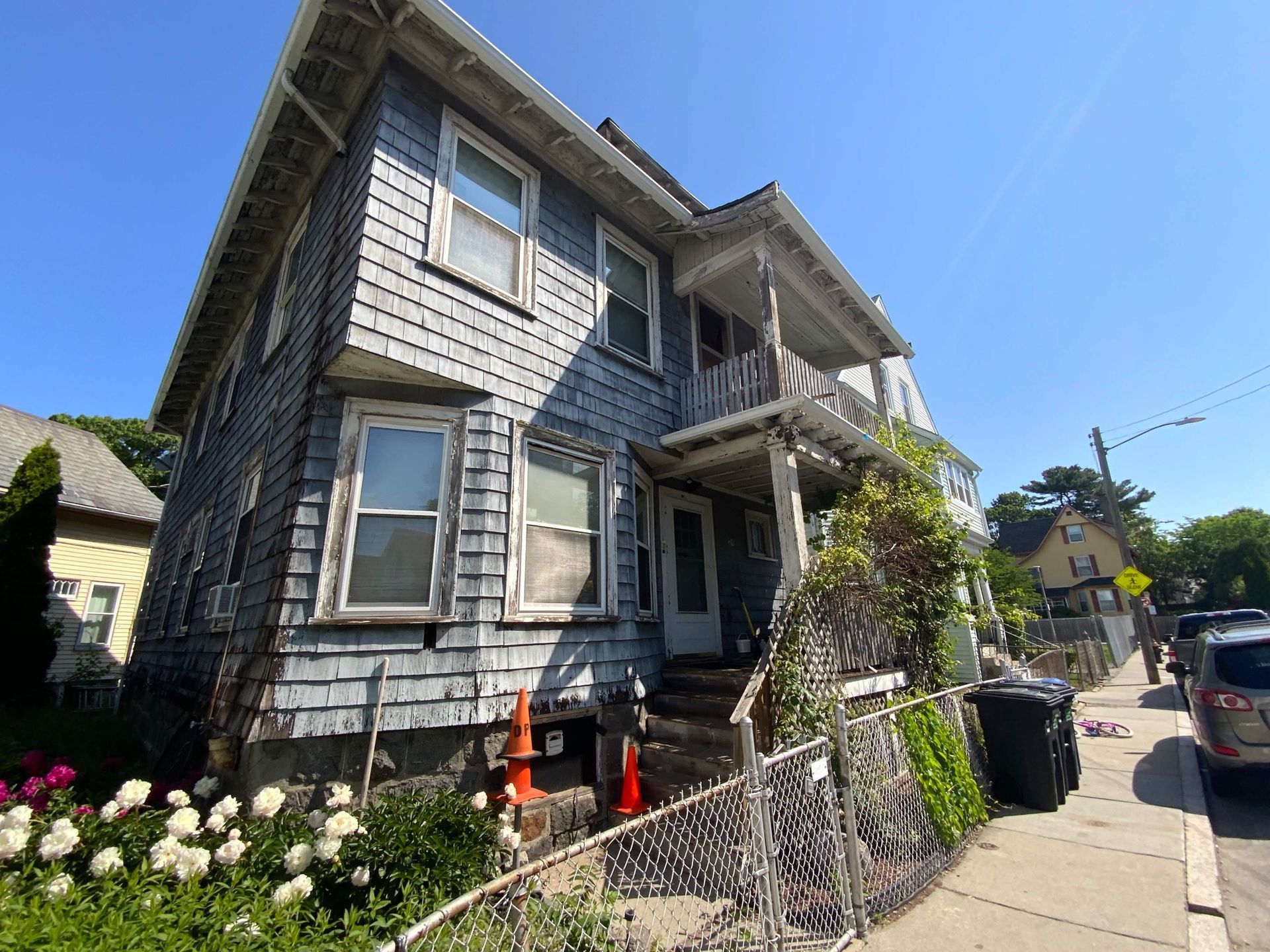 Three-story gray house with weathered shingles, small front porch, and blooming flowers in front.