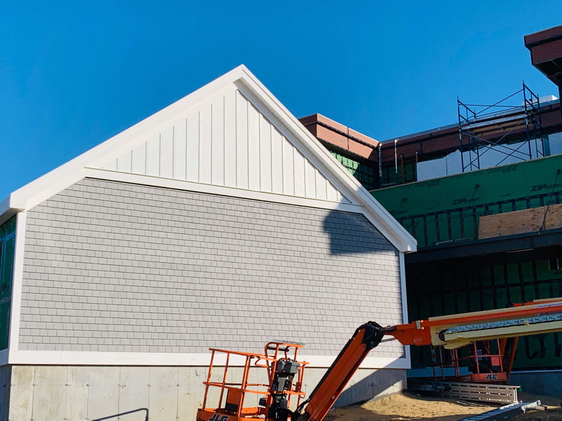 Construction of a building with gray siding and white trim under a blue sky, with an orange lift in the foreground.