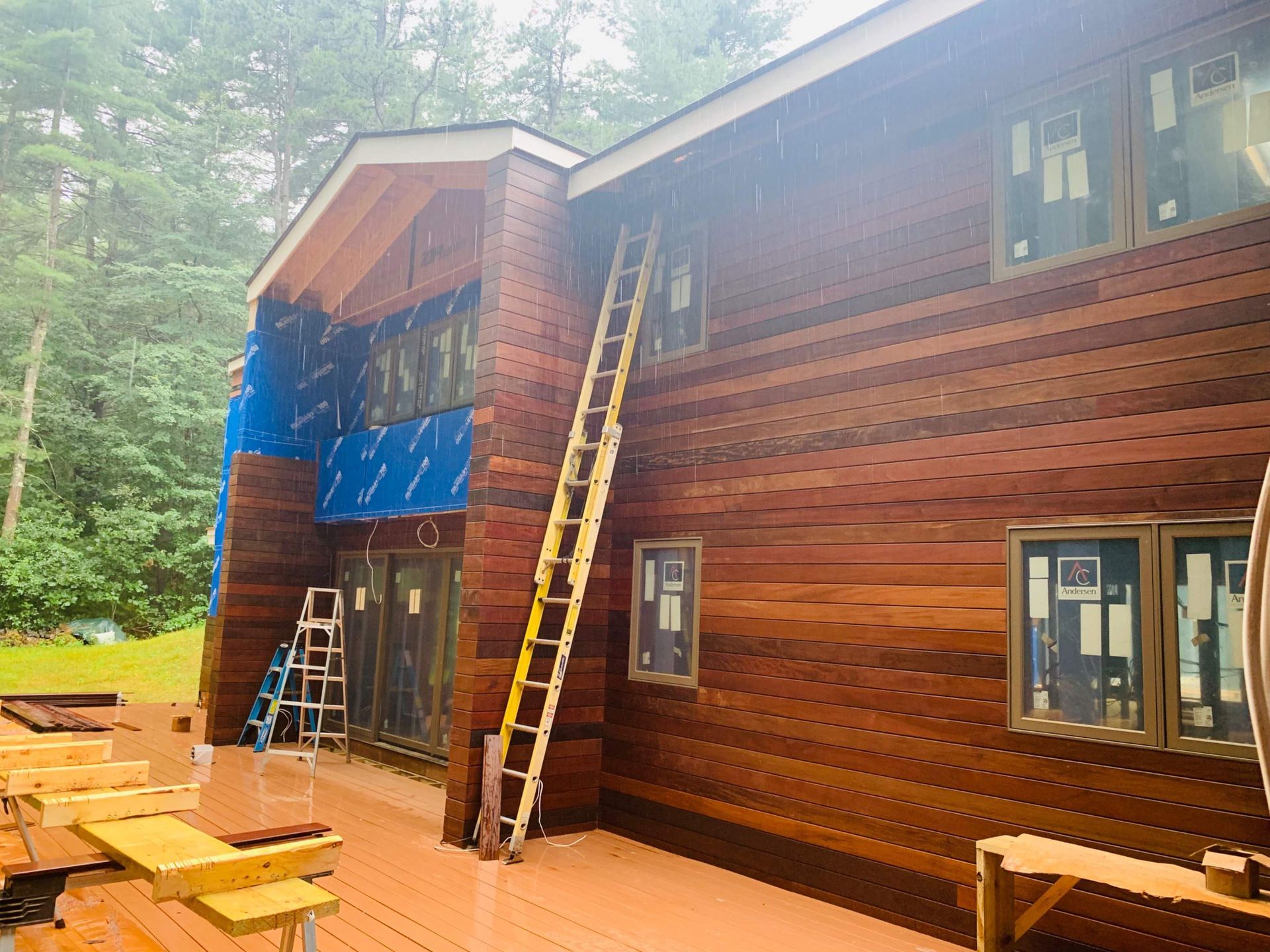 Building exterior with cedar siding, windows, and a ladder. Blue wrap visible. Overcast, rainy day.