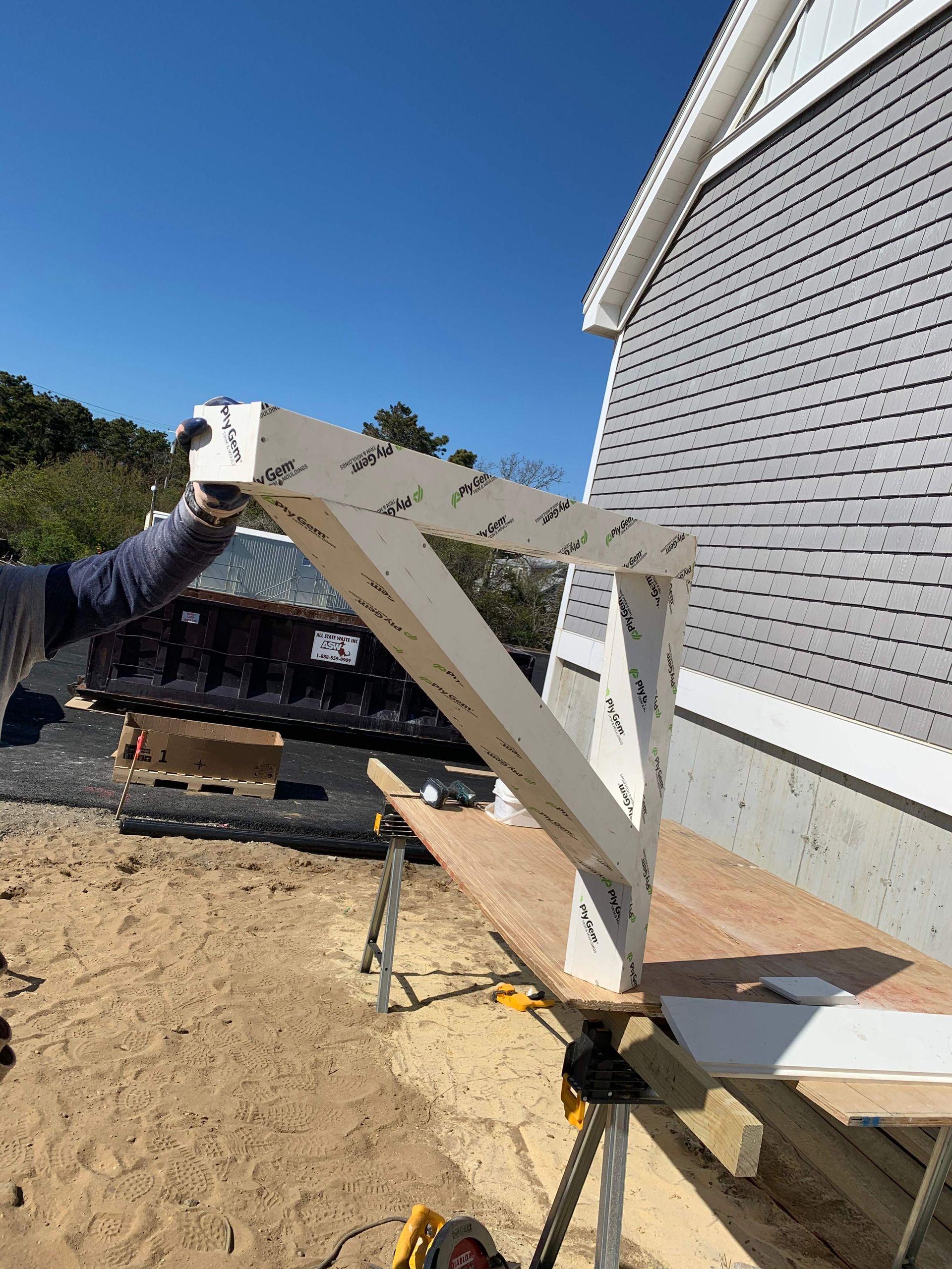 A person holding a partially constructed white, angled structure near a building with gray siding and a blue sky backdrop.