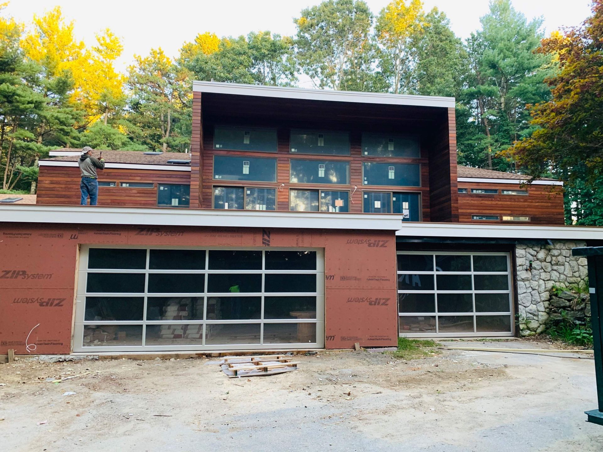 Modern home under construction with large windows and garage doors. A person stands on the roof.