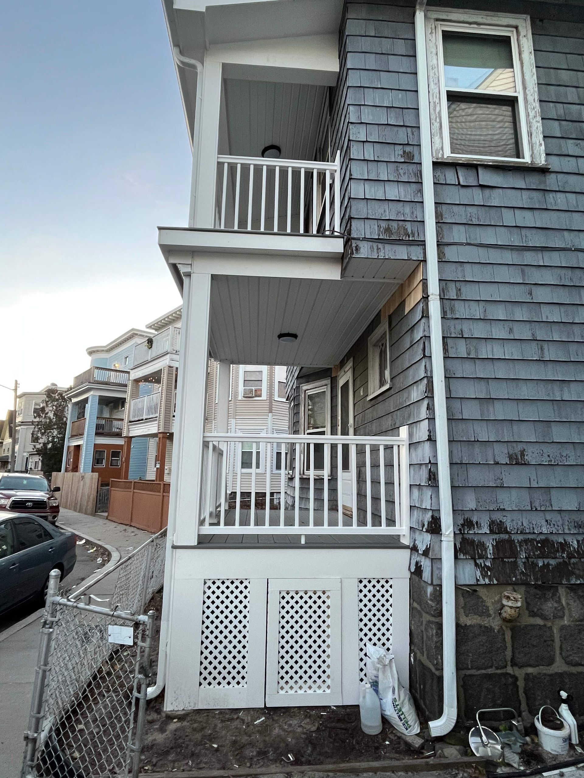 Two-story building exterior with porch and blue siding. White railings and doors. Street with cars and sidewalk.