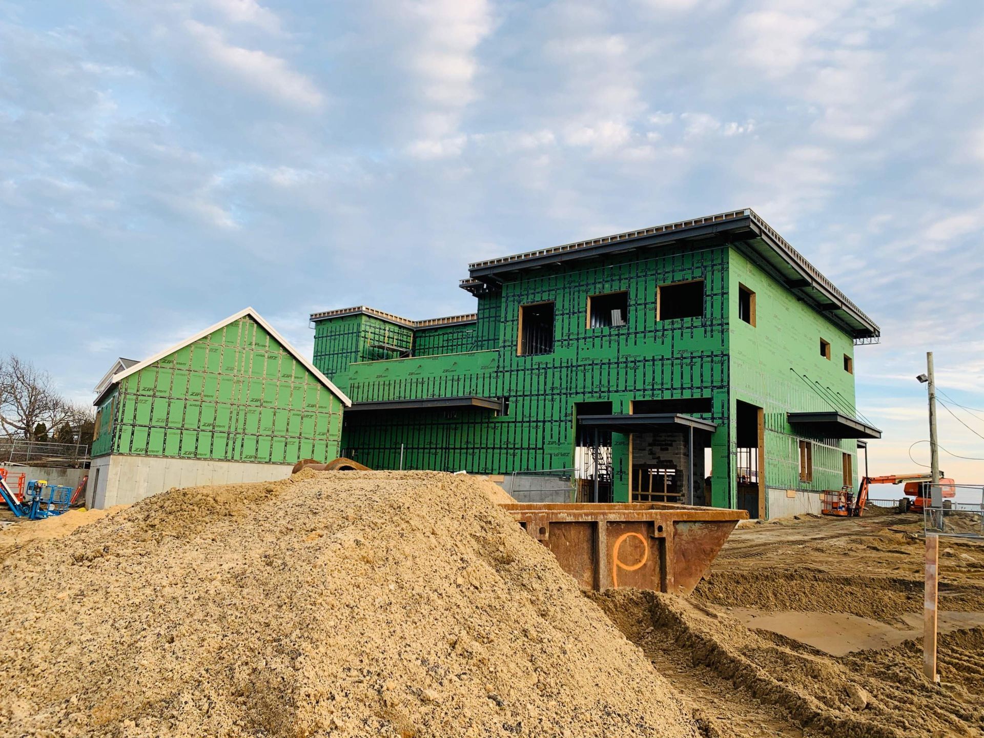 House under construction; green siding, unfinished windows, large pile of dirt in foreground, blue sky.
